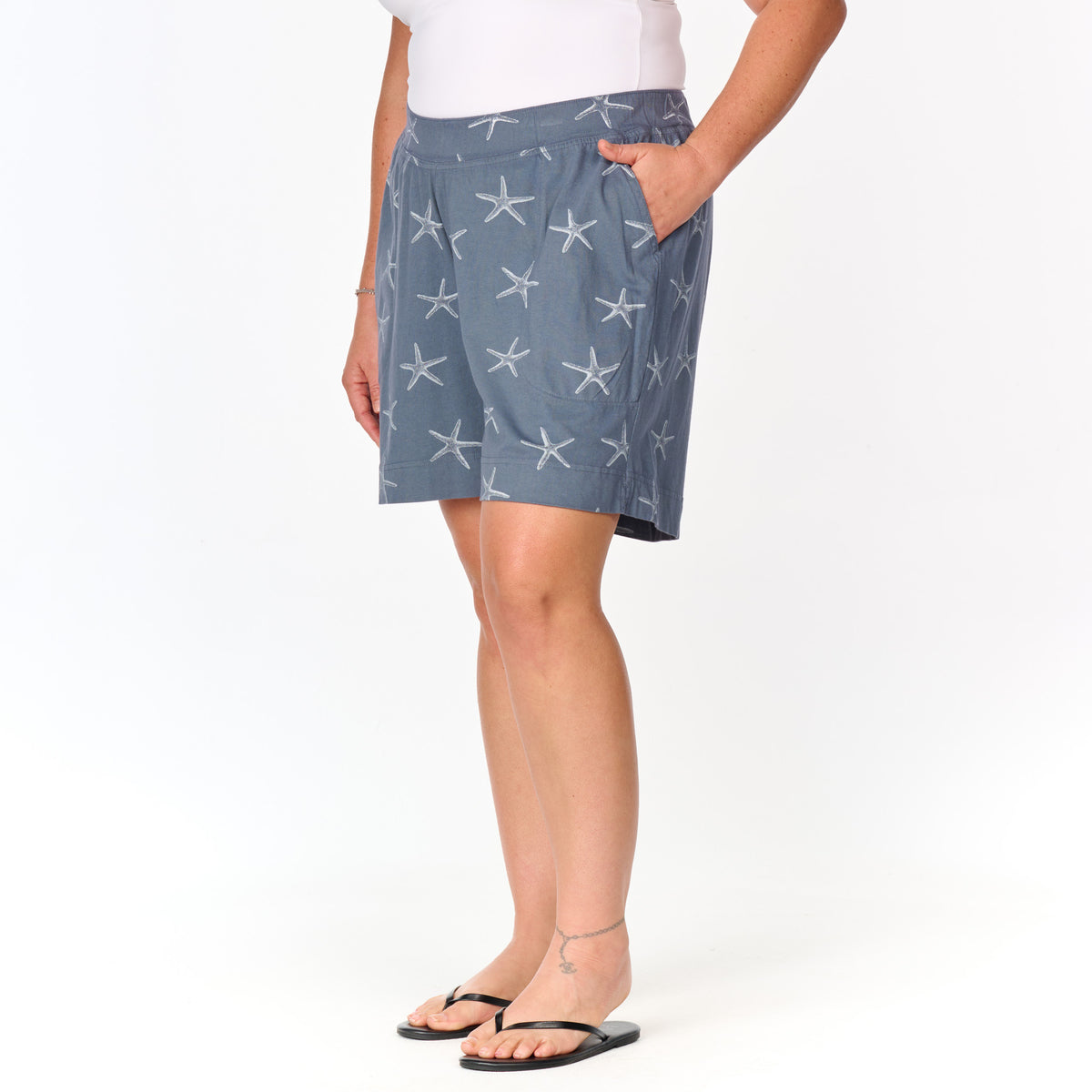 A person wearing a white top and Fresh Produce Clothes’ Sea Star 7″ Key Largo Shorts in plus size, featuring a blue fabric with a white starfish pattern, stands in black flip-flops with one hand in their pocket.
