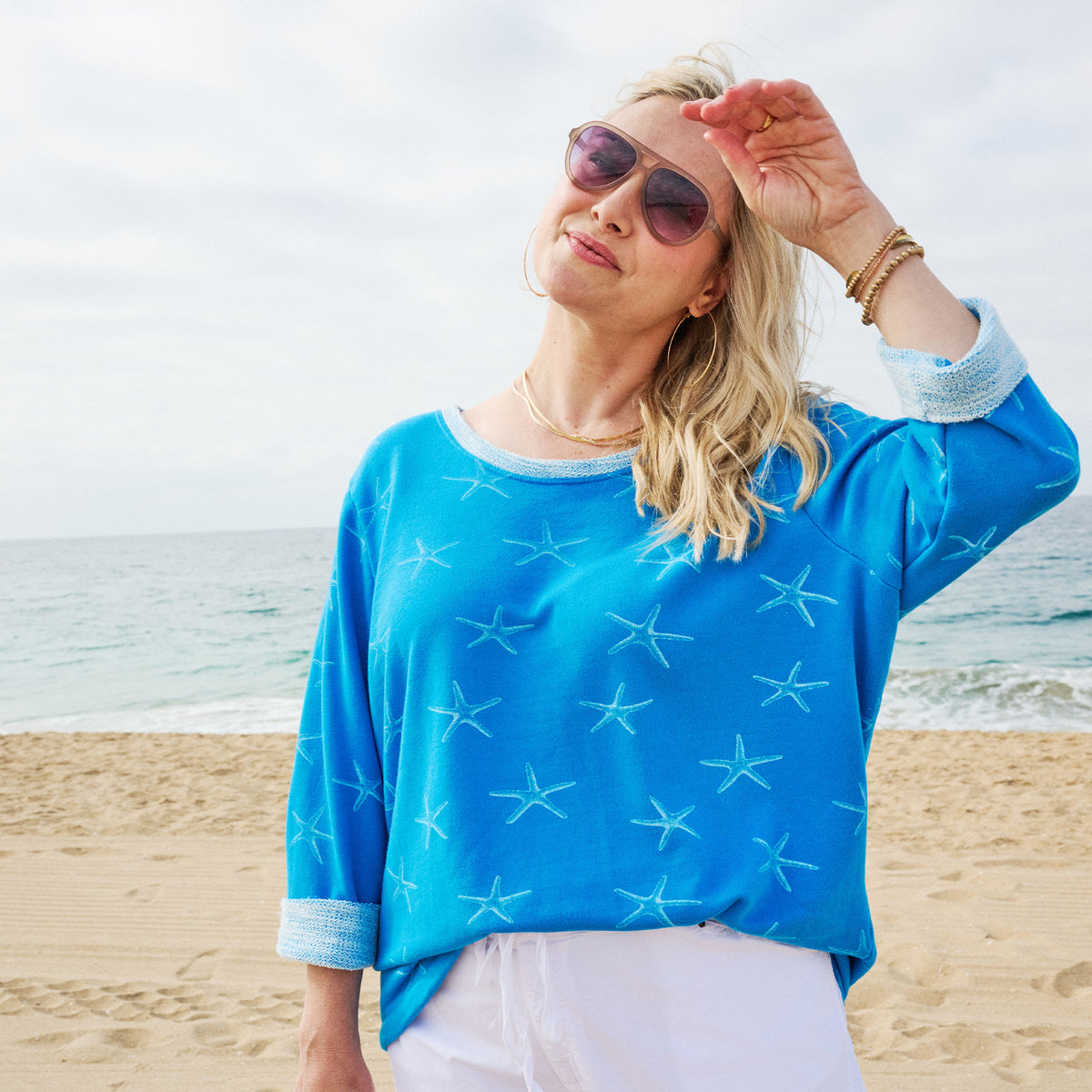 A woman wearing the Fresh Produce Clothes Sea Star Sunset Sweatshirt in plus size, featuring a blue starfish pattern and crafted from soft French terry fabric, stands on a sandy beach shielding her eyes from the sun. The ocean and a partly cloudy sky form the background.