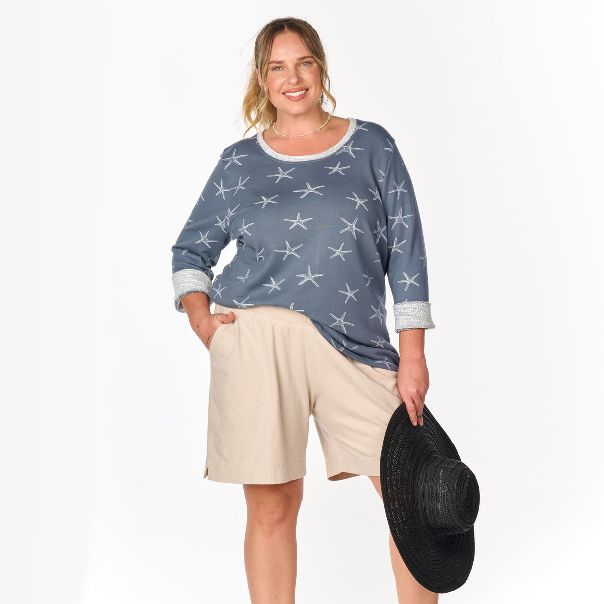 A smiling woman poses against a white background wearing the Fresh Produce Clothes Sea Star Sunset Sweatshirt in plus size and beige shorts, holding a black wide-brimmed hat.