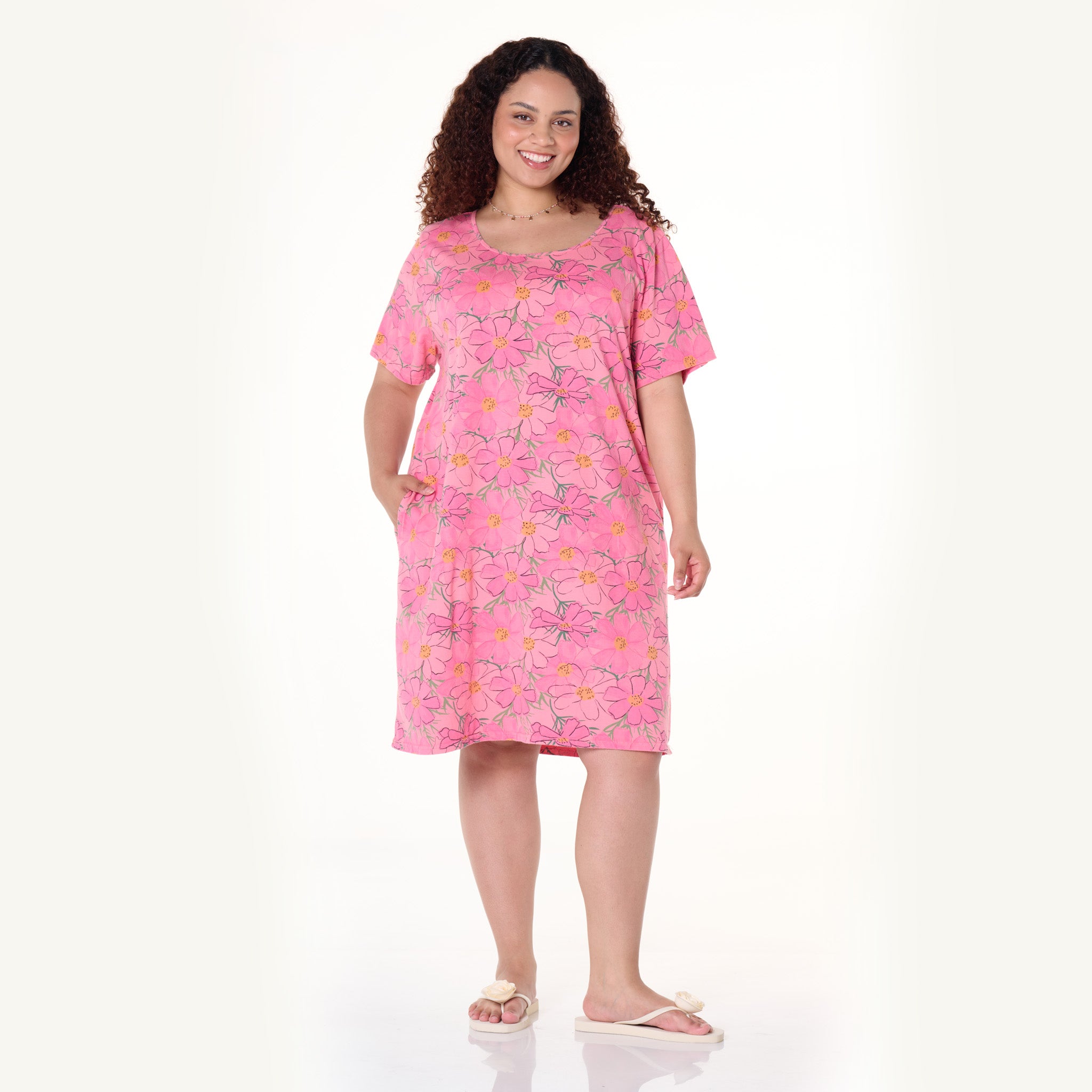 A woman with curly hair stands smiling against a plain white background, wearing the Fresh Produce Clothes Spring Day Sanibel Dress | Plus Size—a comfortable, knee-length pink floral print dress featuring short sleeves and pockets—paired with light-colored slippers.