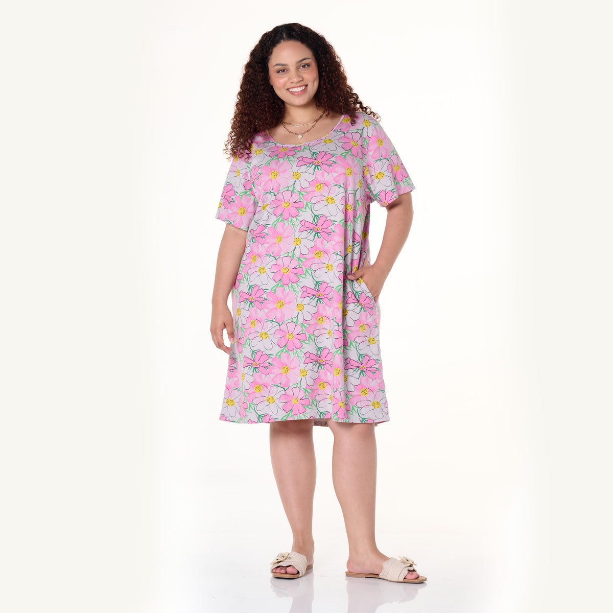 Smiling woman with curly hair wearing the Fresh Produce Clothes Spring Day Sanibel Dress in plus size and sandals, standing with one hand in her pocket against a plain, light background.