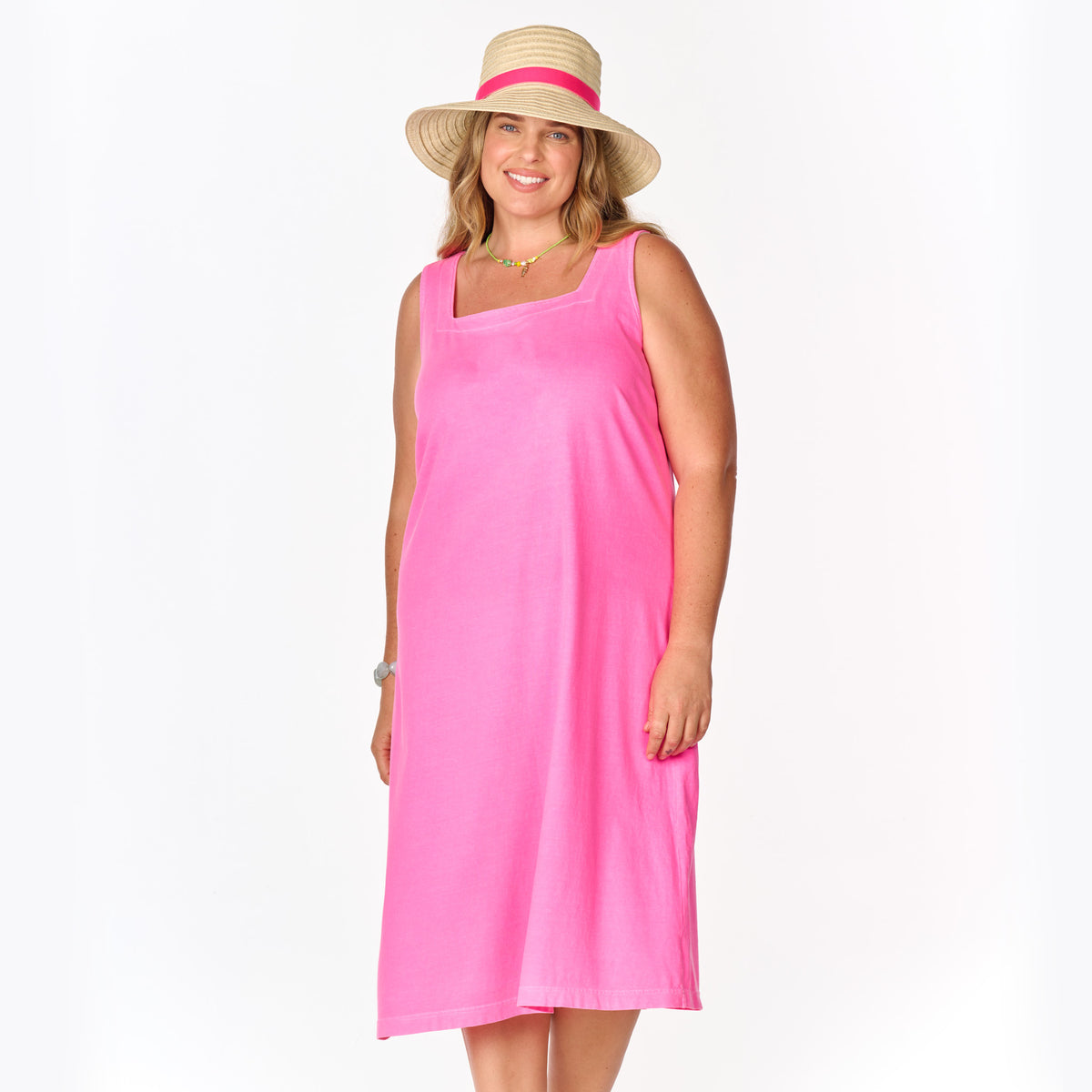 A woman wearing the Fresh Produce Clothes Square Neck Dress in bright pink cotton jersey, paired with a wide-brimmed straw hat with a pink ribbon, smiles while standing against a plain white background.
