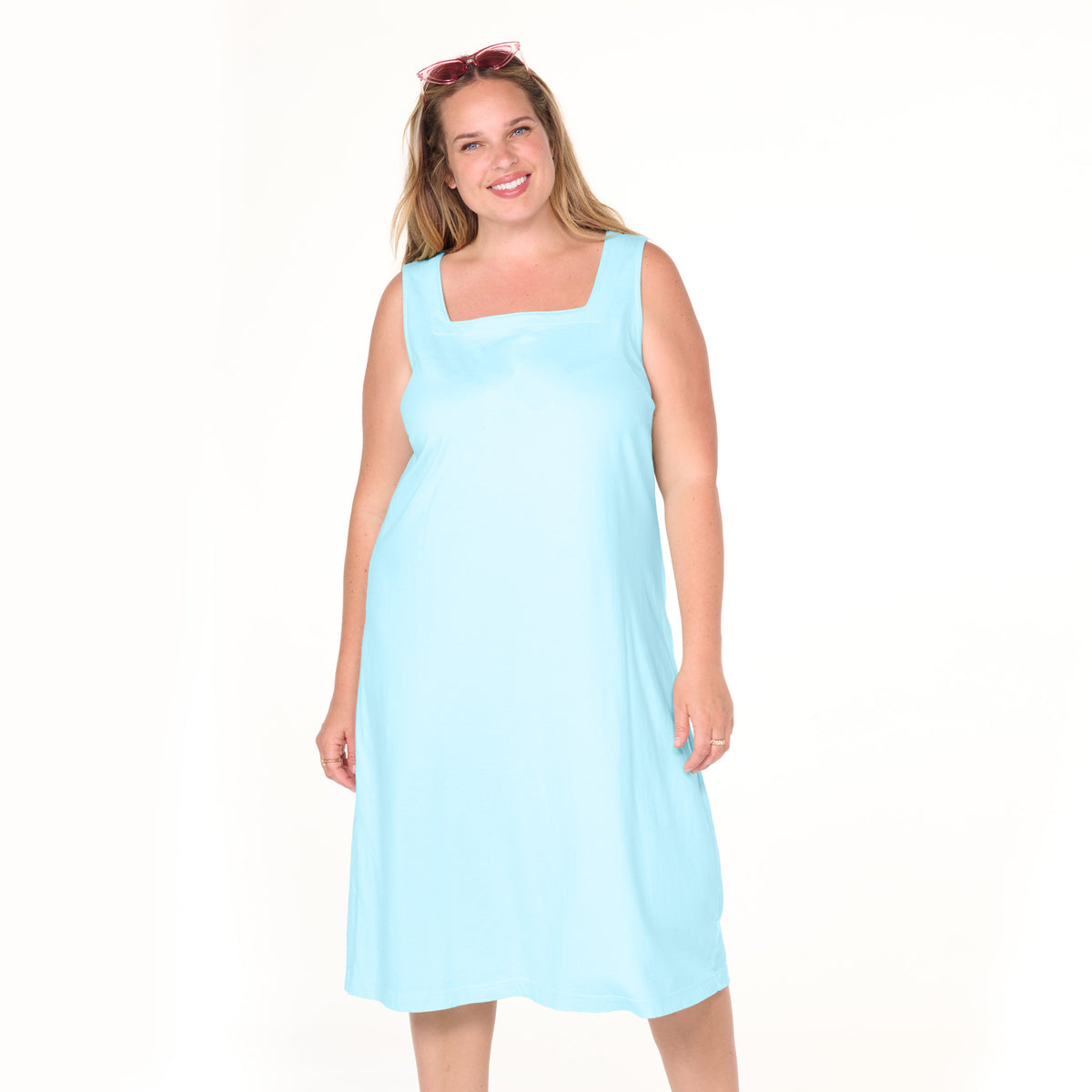 A smiling woman stands against a plain white background, wearing the Fresh Produce Clothes Square Neck Dress | Plus Size—a sleeveless, light blue dress crafted from soft cotton jersey—with pink sunglasses atop her head.