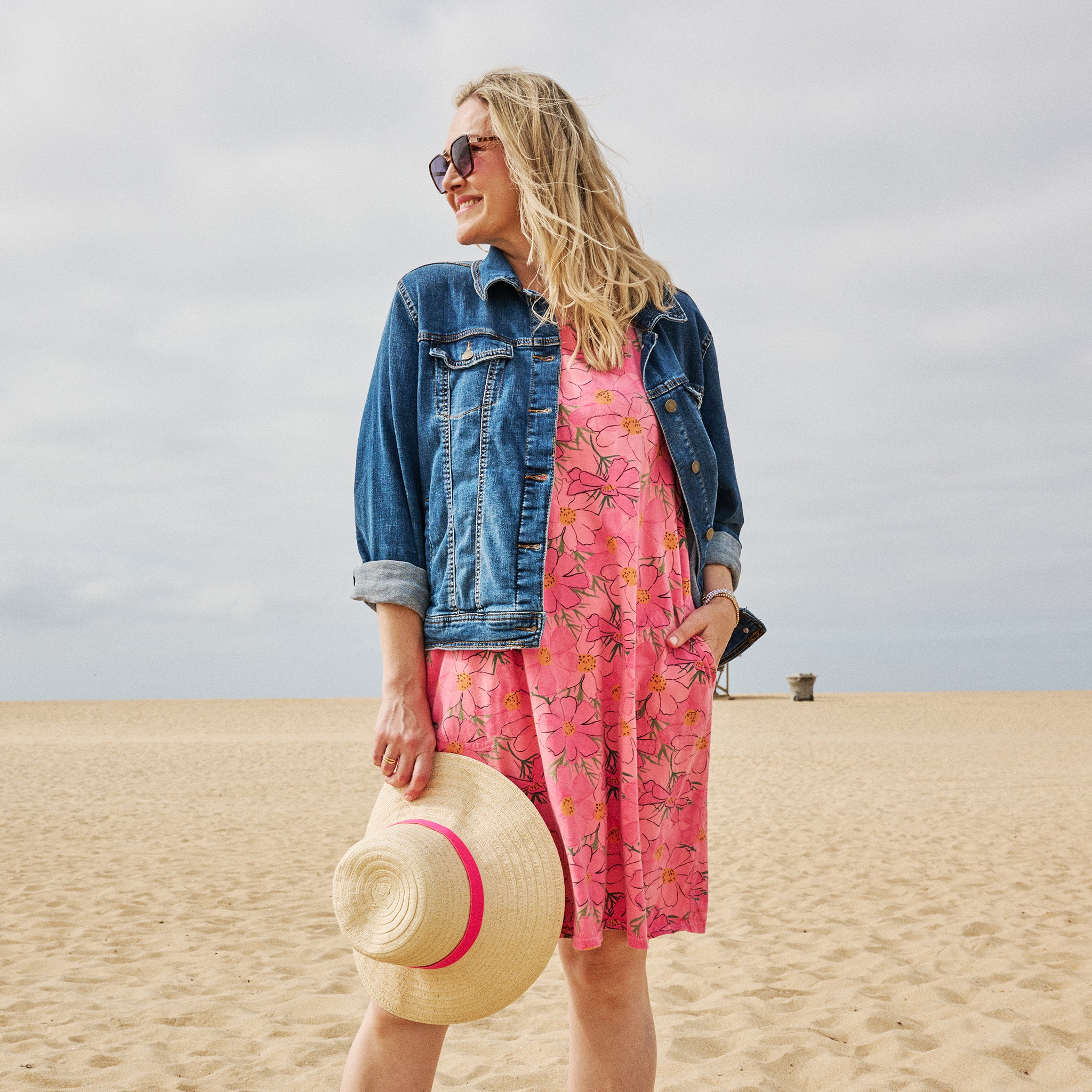 A woman stands on a sandy beach wearing the Spring Day Sanibel Dress by Fresh Produce Clothes in a pink floral print, paired with a denim jacket and sunglasses. She holds a straw hat adorned with a pink ribbon and smiles, looking away from the camera beneath a cloudy sky.
