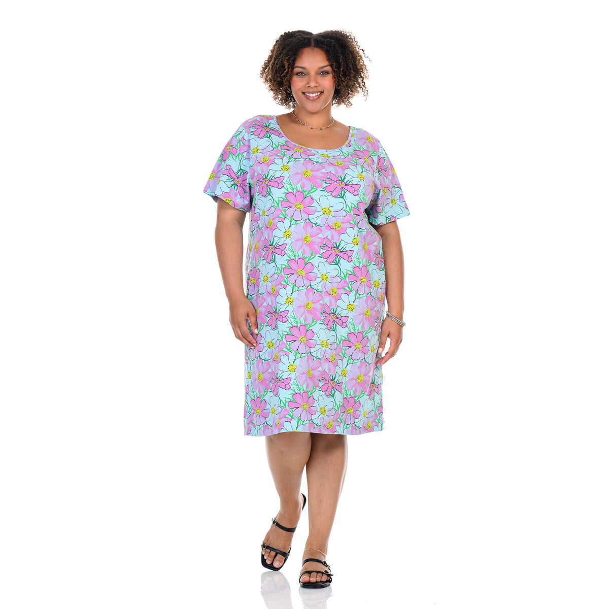 A woman stands smiling against a white background, wearing the Fresh Produce Clothes Spring Day Sanibel Dress | Plus Size—a short-sleeve, knee-length cotton dress with a vibrant floral print featuring pink, green, and blue accents. She pairs the look with black sandals and minimal jewelry. This plus size dress offers both comfort and style for any occasion.
