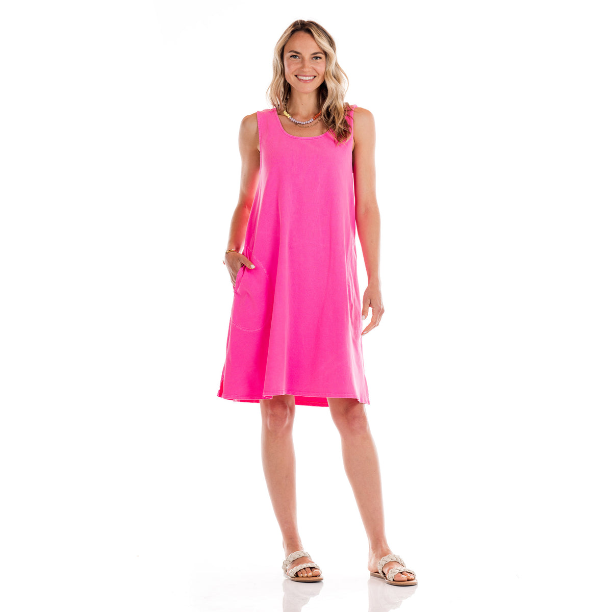 A smiling woman wears the Fresh Produce Clothes Sun Dress, a sleeveless, bright pink cotton jersey style with one hand in her pocket and white sandals. Her wavy hair frames her face as she stands against a plain white background. This effortlessly stylish look is proudly made in the USA.