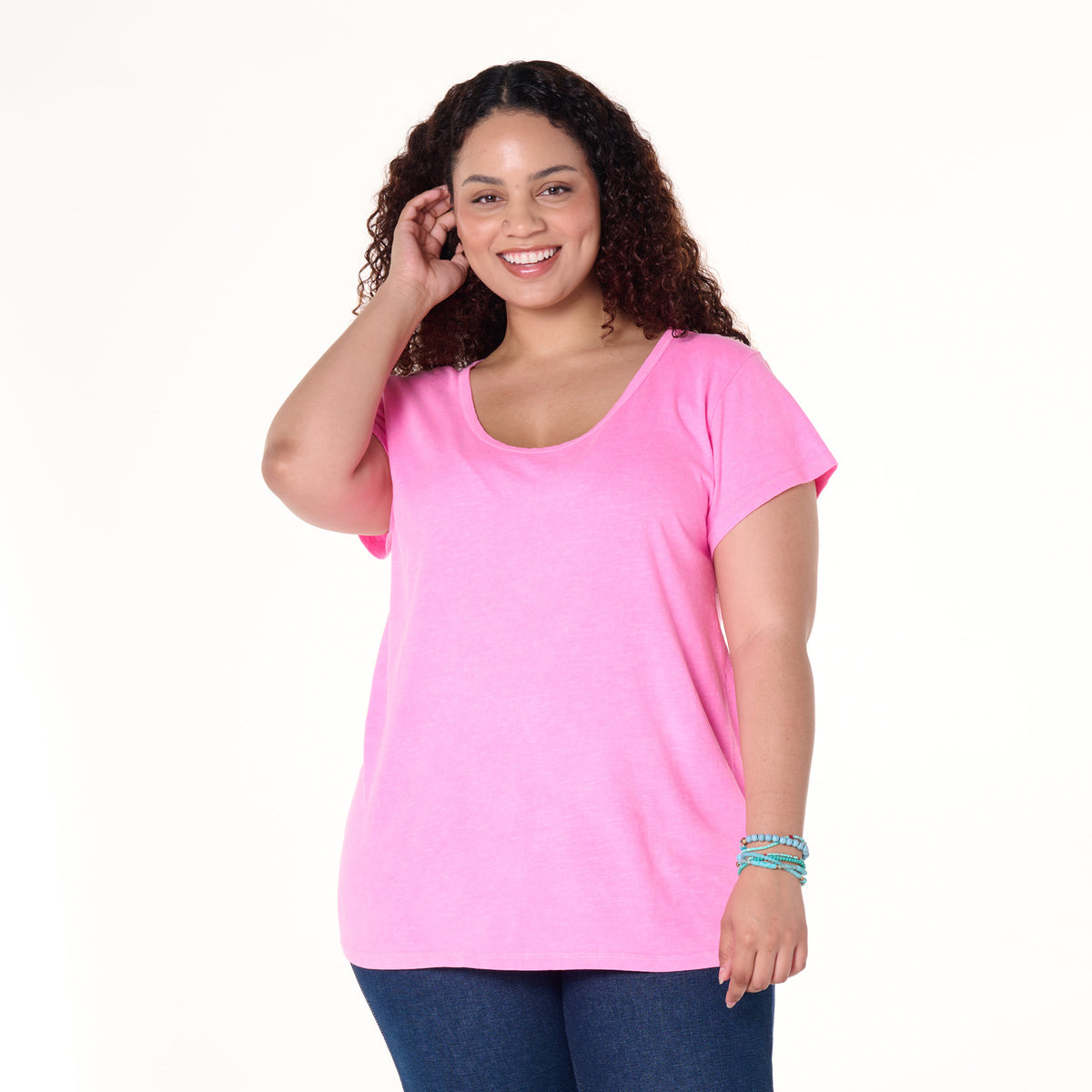 A woman with curly hair smiles, wearing the Fresh Produce Clothes Vintage Scoop Neck T in bright pink and dark blue jeans. She stands against a plain white background, one hand touching her hair and the other by her side, highlighting the slimming shape of this plus size clearance top.