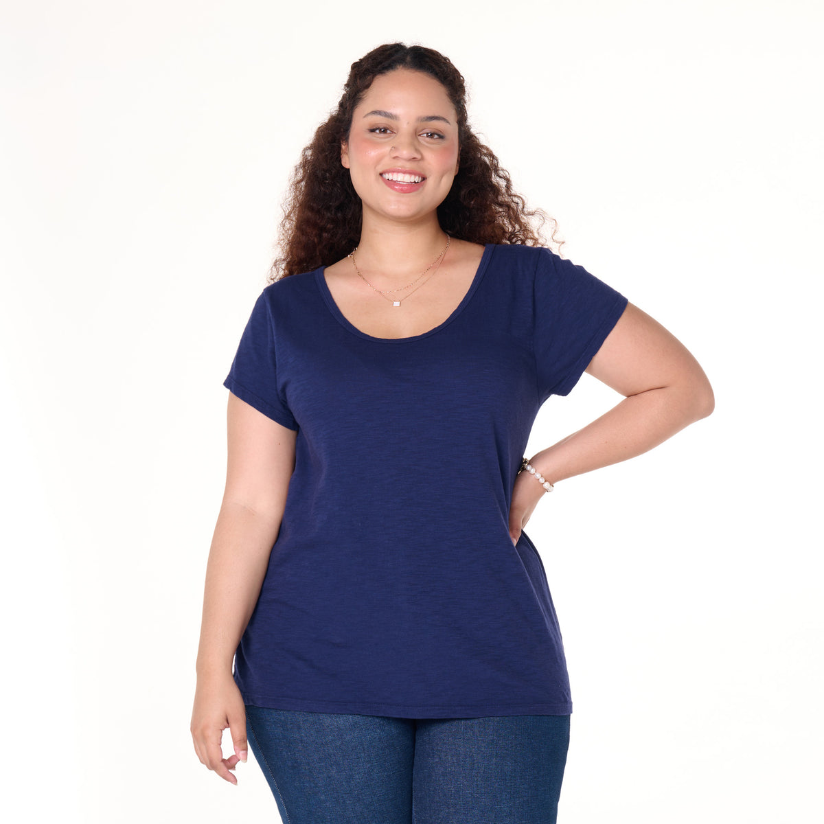 A woman with curly hair, wearing the Fresh Produce Clothes Vintage Scoop Neck T | Plus Size in navy blue and blue jeans, stands smiling with one hand on her hip against a plain white background.