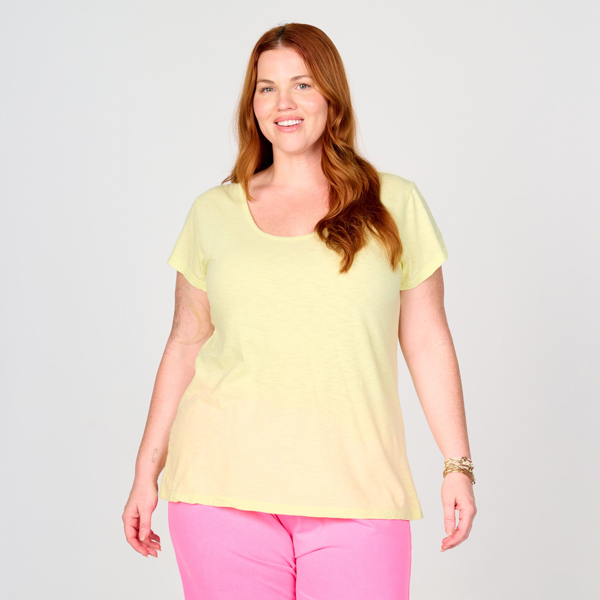 A smiling woman with long red hair is wearing a light yellow Fresh Produce Clothes Vintage Scoop Neck T (Plus Size, Clearance) and bright pink pants, standing against a plain light gray background.