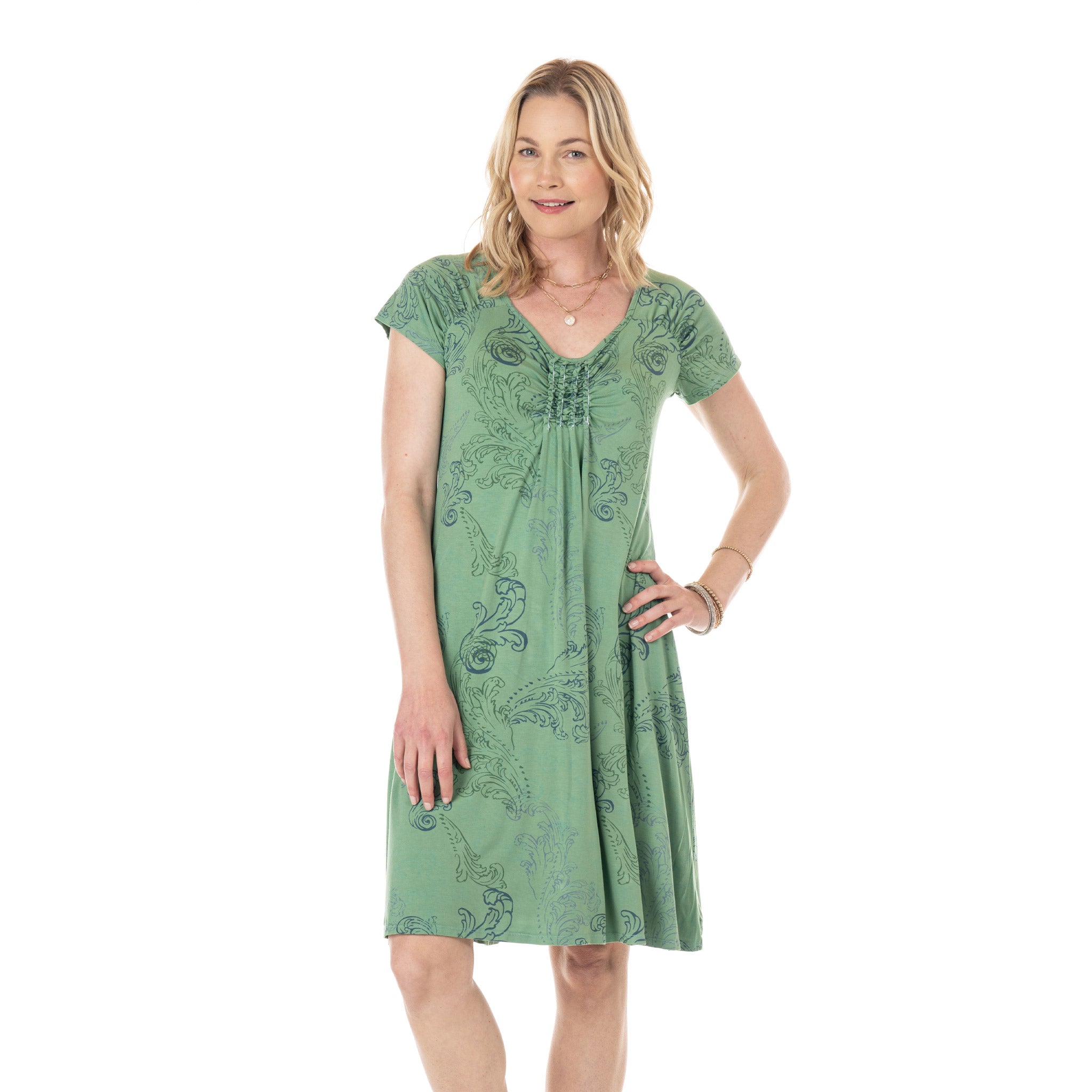 A woman stands smiling with one hand on her hip against a plain white background, wearing the Waverley Swirl Emma Dress Lagoon from Fresh Produce Clothes. This short-sleeve, green patterned A-line dress is crafted from soft rayon lycra for a flattering fit and effortless movement.