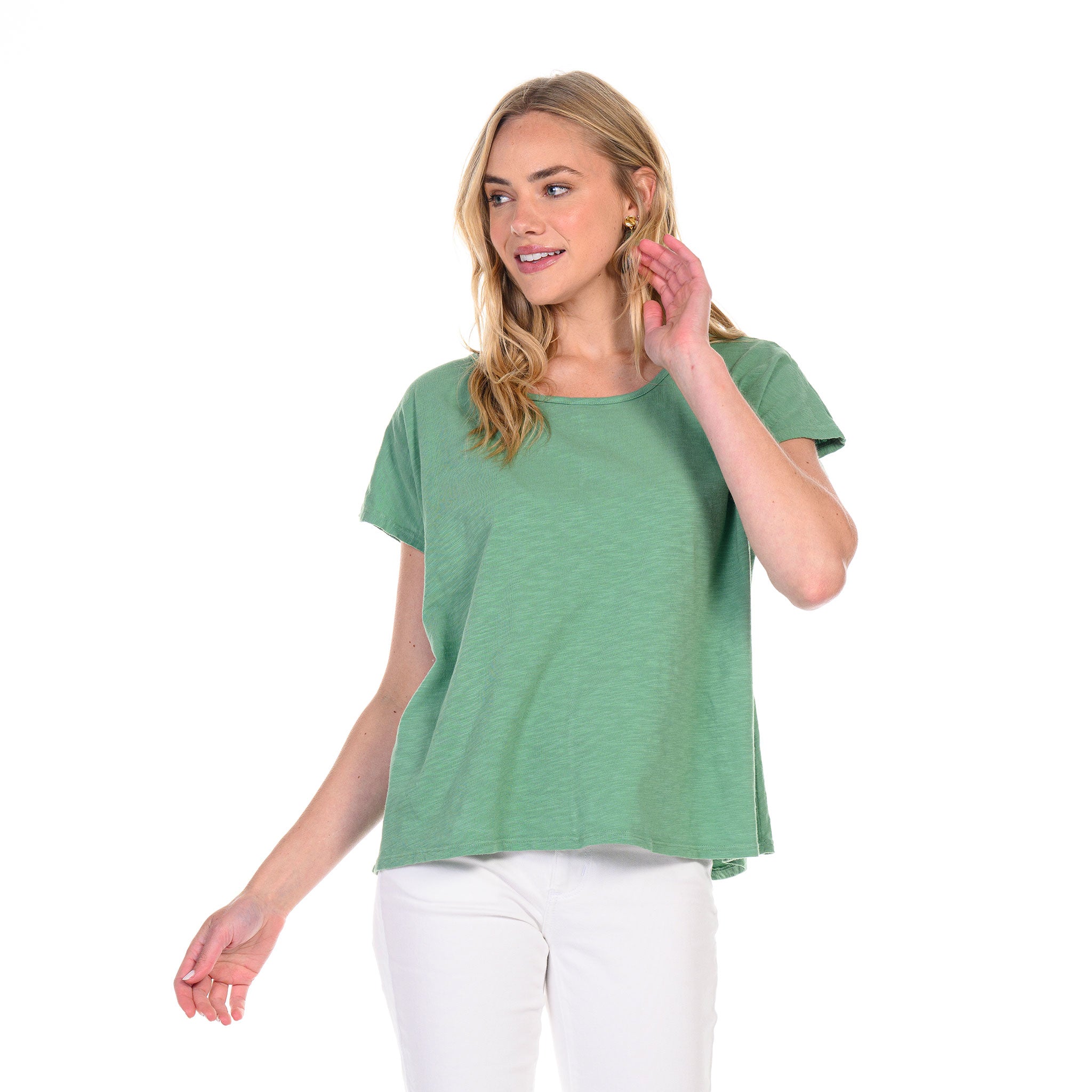 A woman stands against a white background, touching her hair with one hand and looking to the side, wearing a loose, slouchy fit green Fresh Produce Clothes Keepsake T (Clearance) with white pants.