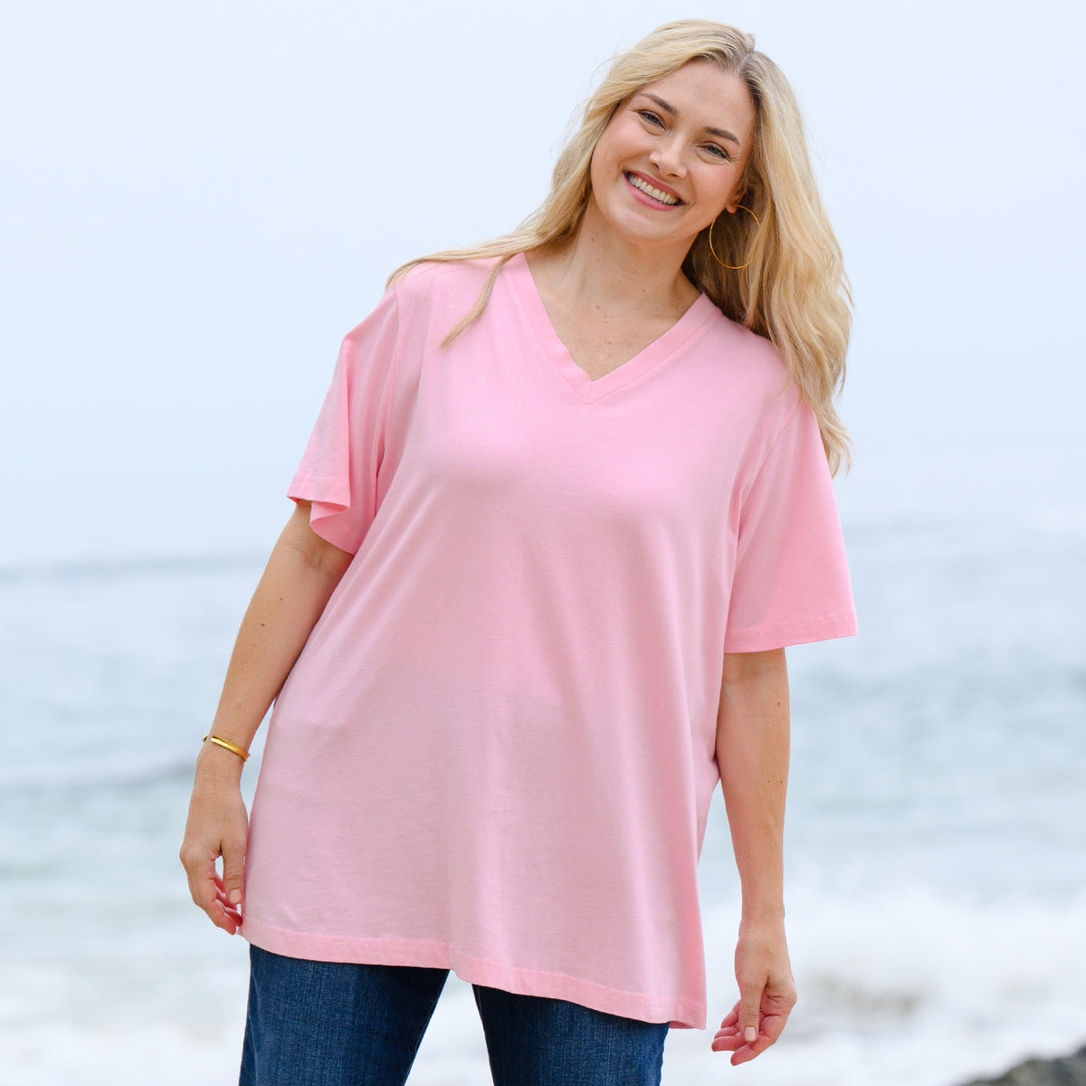 A smiling woman with long blonde hair stands outdoors near the ocean, waves and a cloudy sky in the background. She wears the Fresh Produce Clothes V-Neck T Blush | Plus Size with jeans, enjoying the comfort and style of its relaxed cotton jersey fabric.