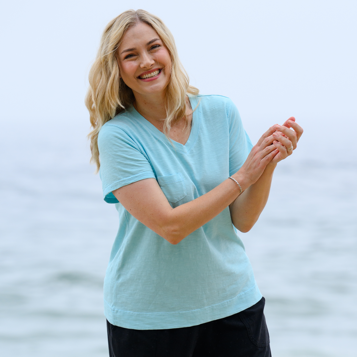 A smiling woman with long blonde hair stands by the water, enjoying a bright outdoor day at the beach in the Heritage Logo Fem T | Plus Size by Fresh Produce Clothes, paired with dark pants.