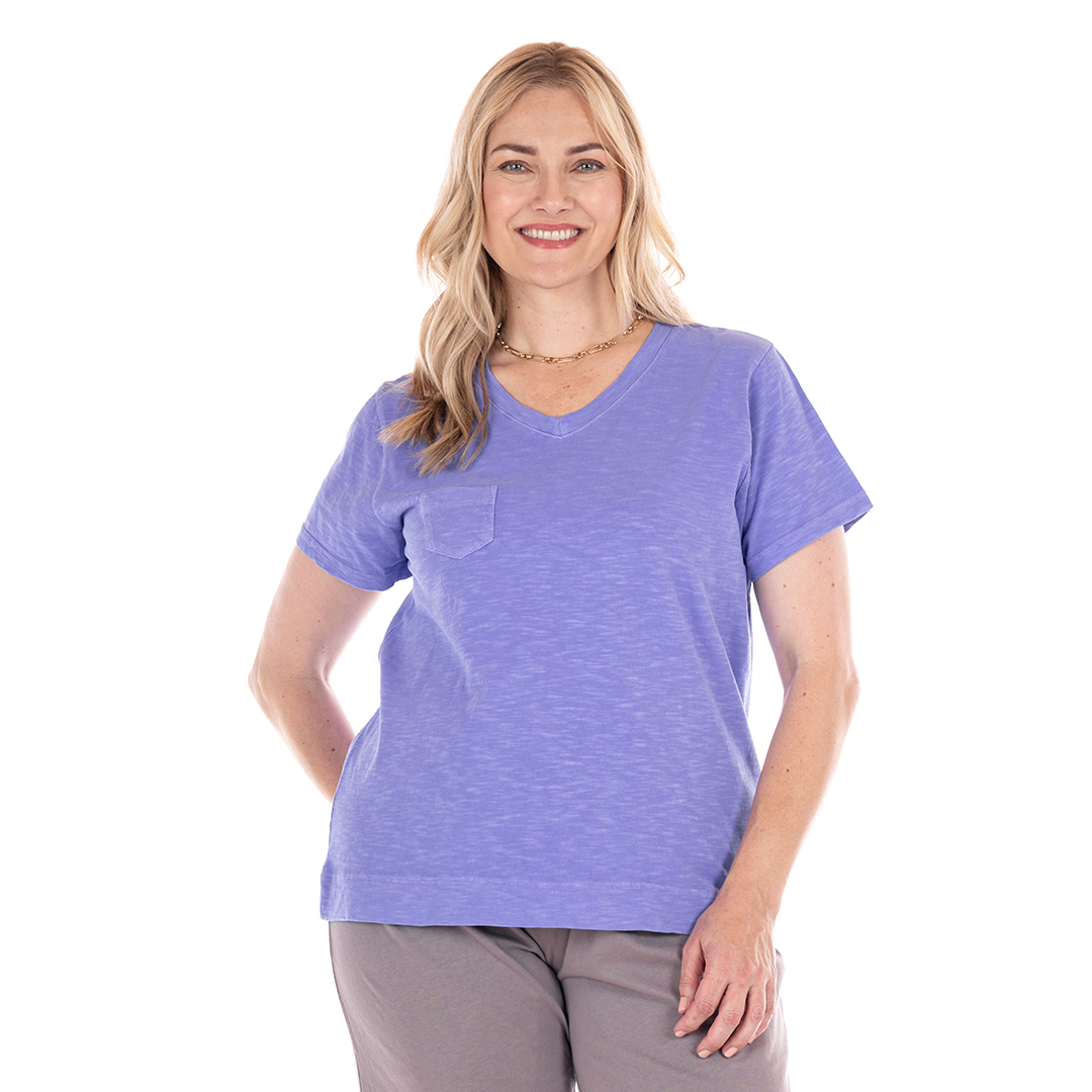 A woman with long blonde hair smiles at the camera. She is wearing a light purple Fresh Produce Clothes Heritage Logo Fem T | Plus Size—a comfortable cotton slub V-neck tee with a pocket—paired with gray pants, standing against a white background.
