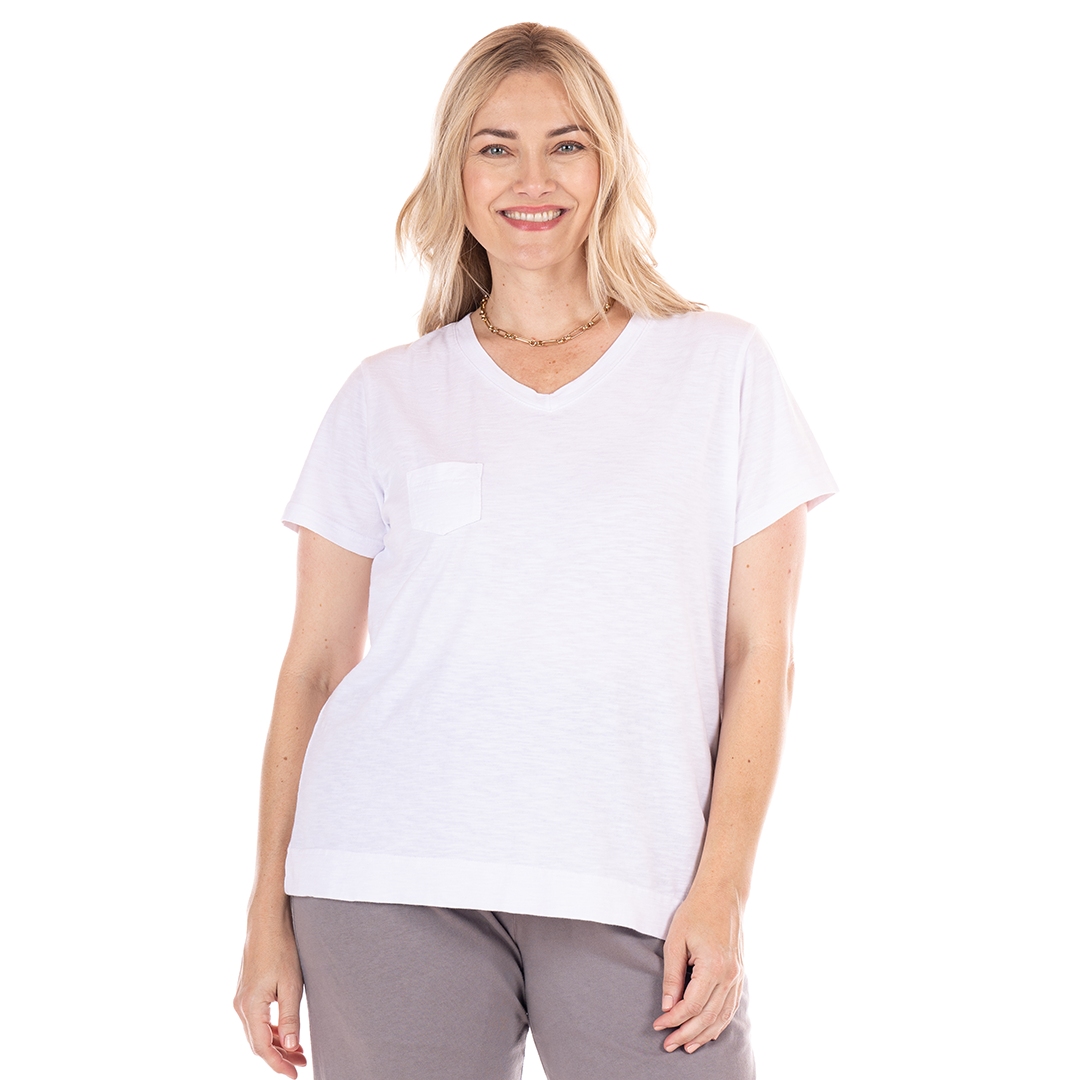A woman with blonde hair smiles while wearing the Fresh Produce Clothes Heritage Logo Fem T | Plus Size in white cotton slub and gray pants, standing against a plain white background.