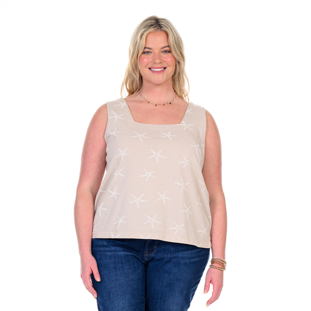 A smiling woman with blonde hair wears the Fresh Produce Clothes Sea Star Boxy Tank in plus size—a sleeveless beige cotton top with a starfish pattern—paired with blue jeans, standing against a plain white background.