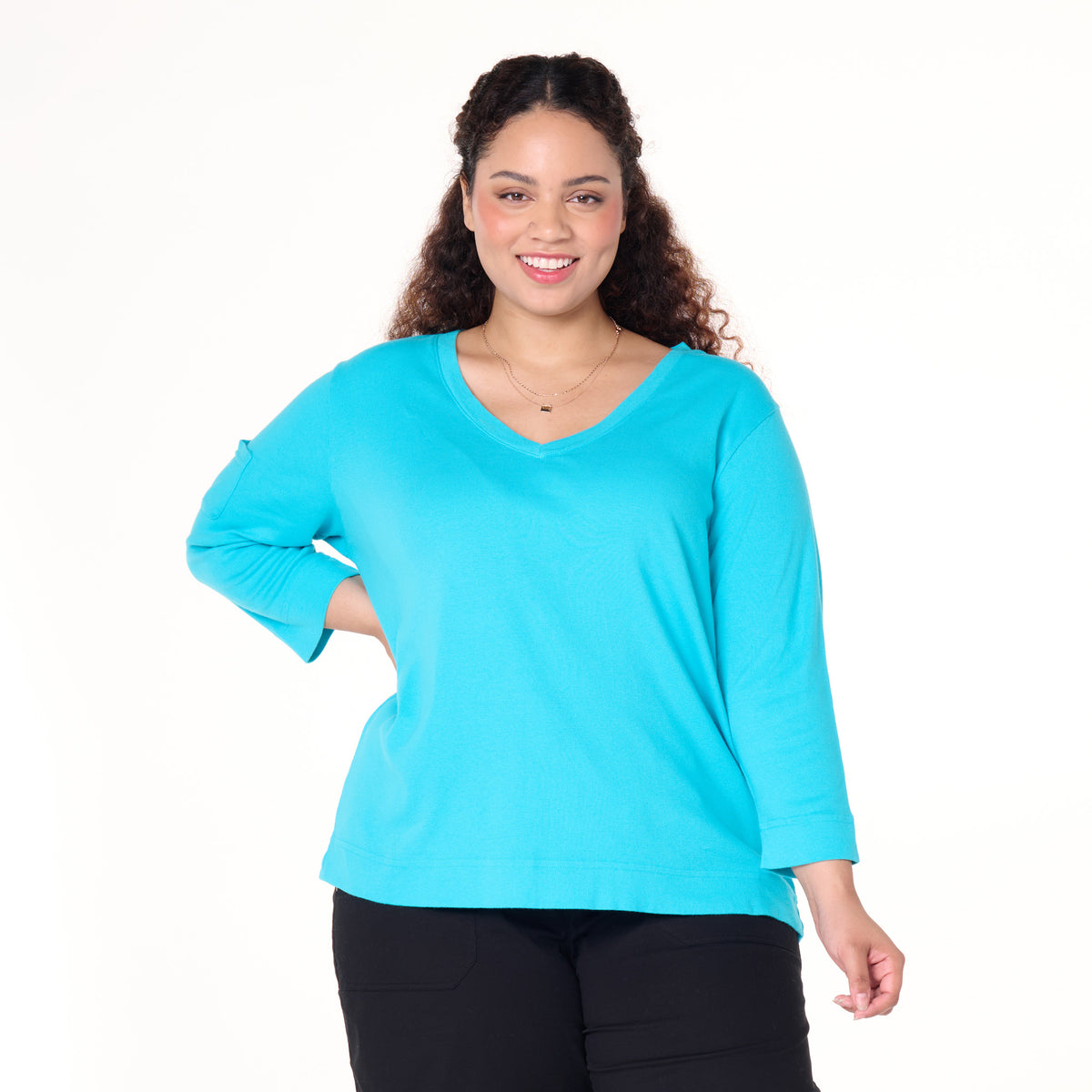 A woman with curly hair is smiling and standing against a plain white background, wearing the Fresh Produce Clothes 3/4 Sleeve Logo Fem T in plus size (Clearance)—a bright turquoise V-neck cotton tee—paired with black pants.