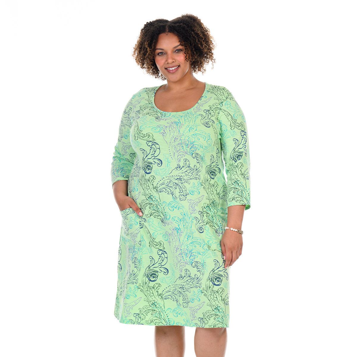 A woman with curly hair smiles and stands against a white background, wearing the Fresh Produce Clothes Waverley Swirl Dalia Dress in plus size. The dress features a light green color with blue Waverley Swirl patterns, 3/4 sleeves, and convenient side pockets—her left hand casually rests in one pocket.