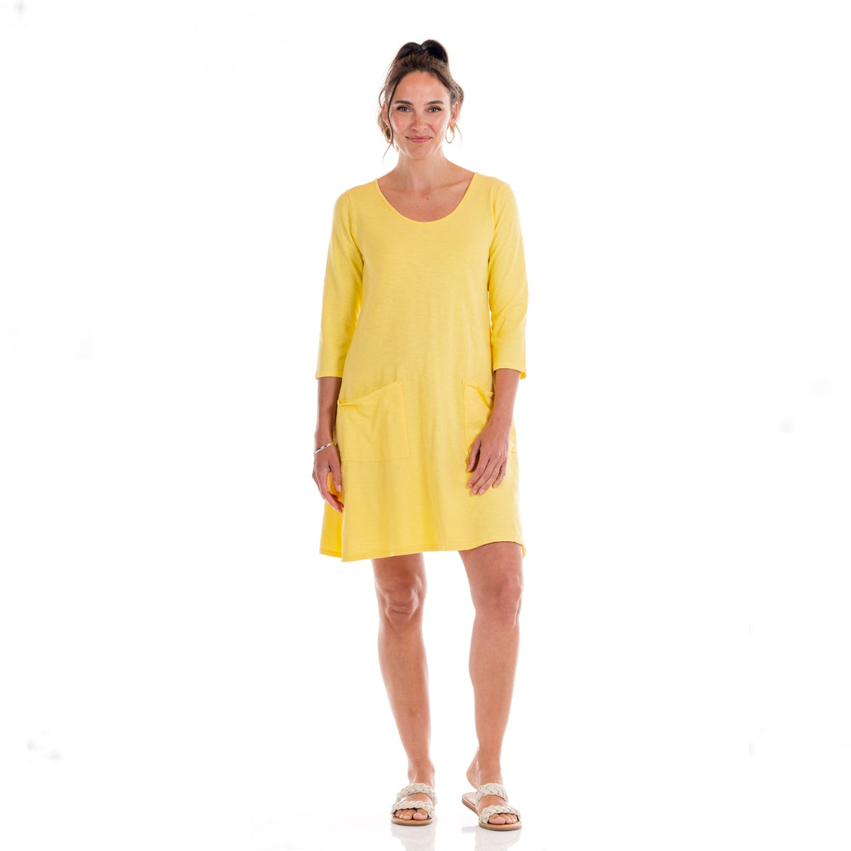 A woman stands smiling in the Dalia Dress Daffodil-Lagoon by Fresh Produce Clothes—a yellow, knee-length cotton slub dress featuring three-quarter sleeves and front pockets—paired with white sandals, against a plain white background. This style is proudly made in the USA.