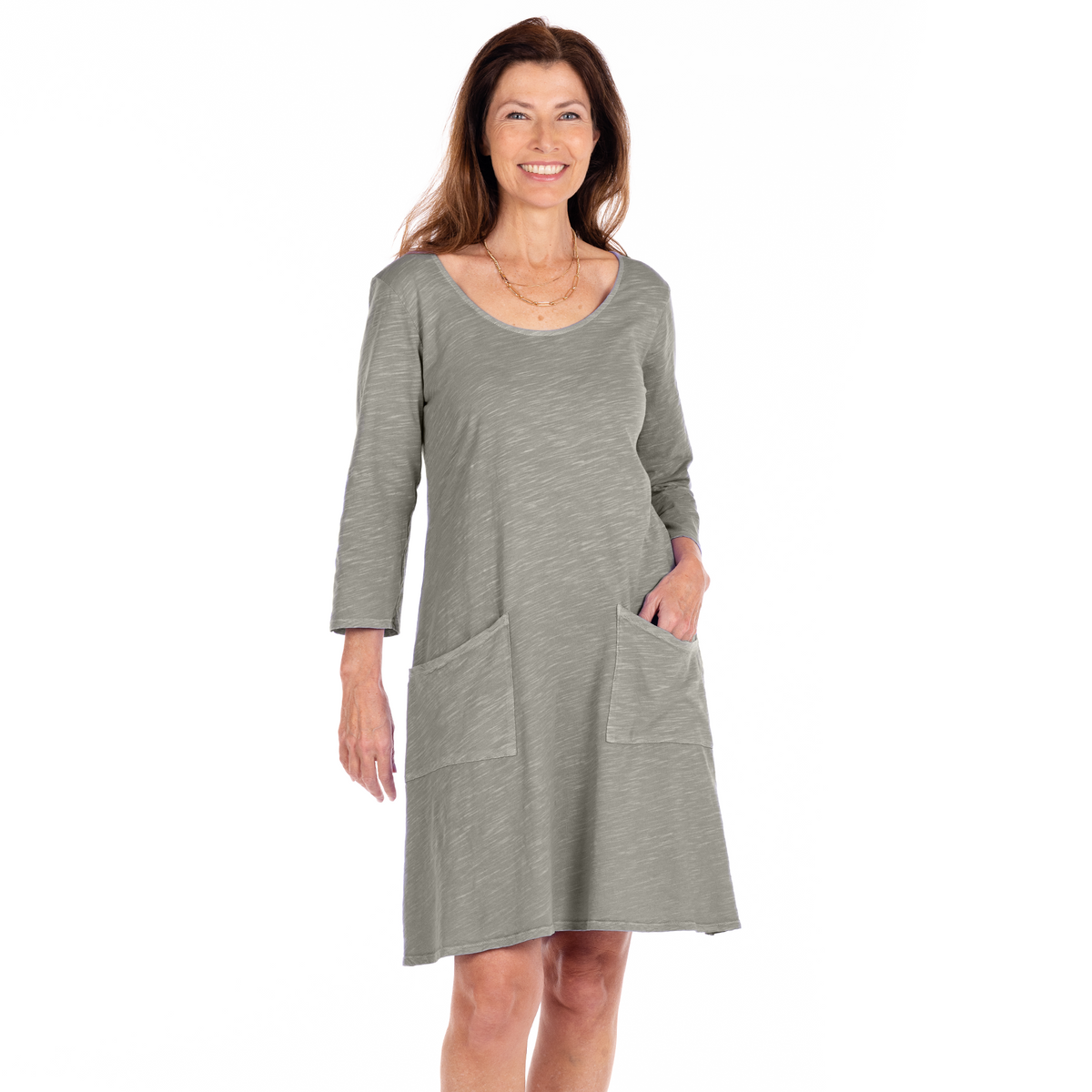 A smiling woman with long brown hair is wearing the versatile Dalia Dress by Fresh Produce Clothes—a light gray, long-sleeve, knee-length dress with two large front pockets—standing against a plain white background. This style is featured in the Clearance section.