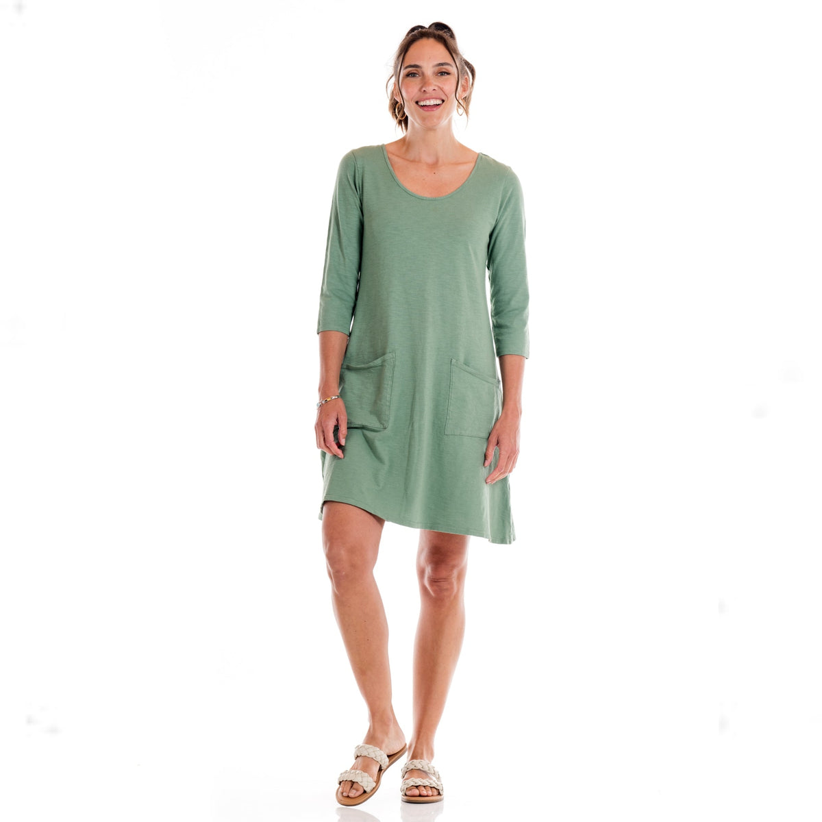 A woman stands smiling in the light green Dalia Dress Daffodil-Lagoon by Fresh Produce Clothes, a knee-length cotton slub dress featuring three-quarter sleeves and front pockets. She wears sandals and has her hair tied back, standing against a plain white background. This stylish look is proudly made in the USA.