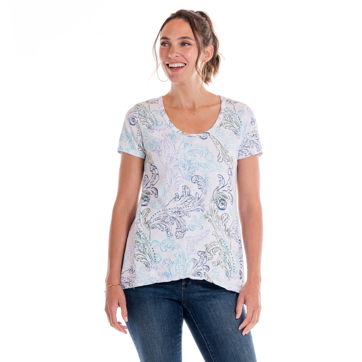 A smiling woman with brown hair in a ponytail wears the Fresh Produce Clothes Waverley Swirl Luna Top | Plus Size, a flowy short-sleeve tee made from soft cotton slub featuring a light-colored paisley pattern, paired with blue jeans and standing against a white background.