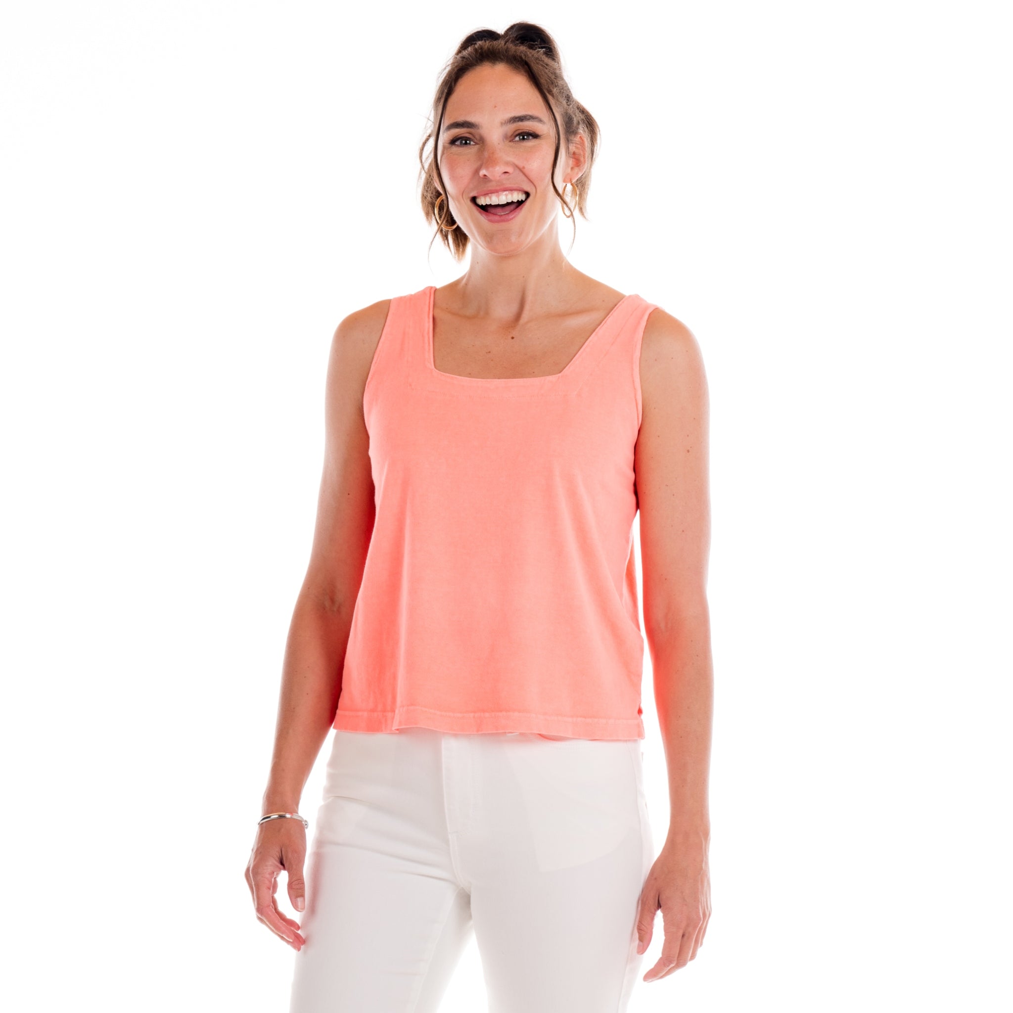 A woman smiles while standing against a white background, wearing the Fresh Produce Clothes Boxy Tank in Sunset Coral—a sleeveless square neck top crafted from soft cotton jersey—and white pants.