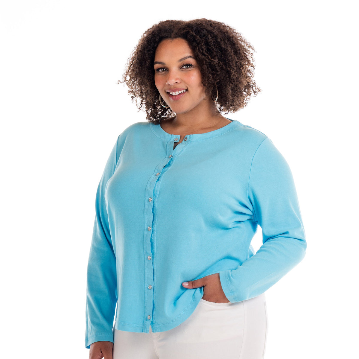 A woman wearing the Fresh Produce Clothes Baby Rib Cardigan | Plus Size BFN CHB in light blue with pearl snaps and white pants smiles at the camera, standing with one hand in her pocket against a white background.