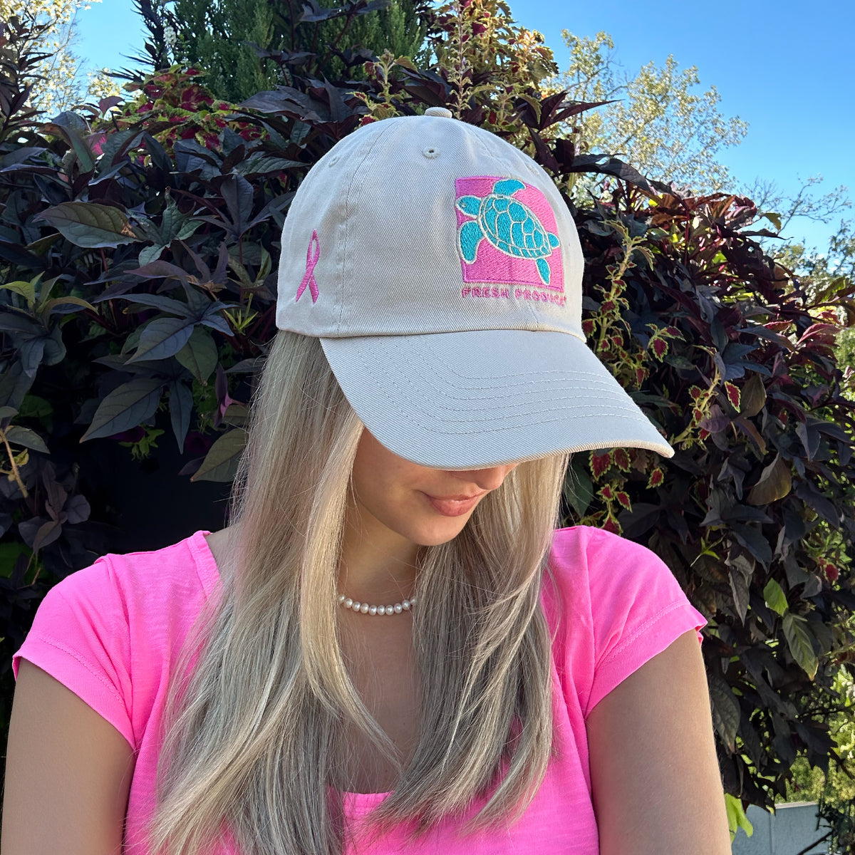 A woman with long blonde hair, wearing a pink shirt and the Breast Cancer Turtle Hat from Fresh Produce Clothes, stands outdoors in front of green foliage.