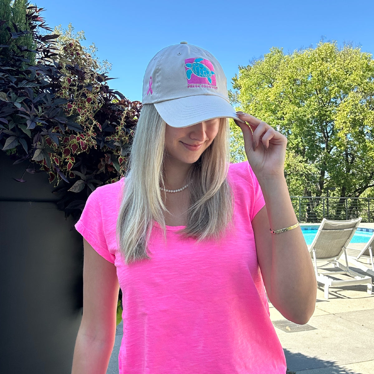 A woman in a bright pink shirt stands by a pool on a sunny day, smiling and holding her Breast Cancer Turtle Hat by Fresh Produce Clothes. Trees and patio furniture are visible in the background.