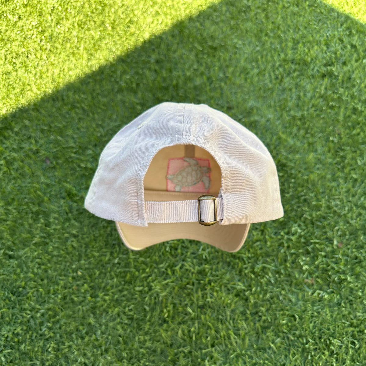 A Breast Cancer Turtle Hat by Fresh Produce Clothes is upside down on green grass, revealing the back adjustable strap and inner tag with sunlight casting a shadow over the scene.