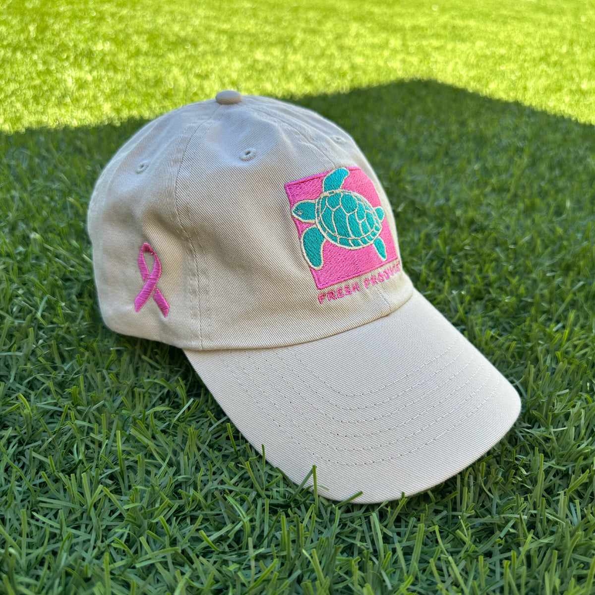 The Breast Cancer Turtle Hat by Fresh Produce Clothes, beige with a pink ribbon on the side and a turquoise sea turtle with a pink background on the front, sits on green grass.