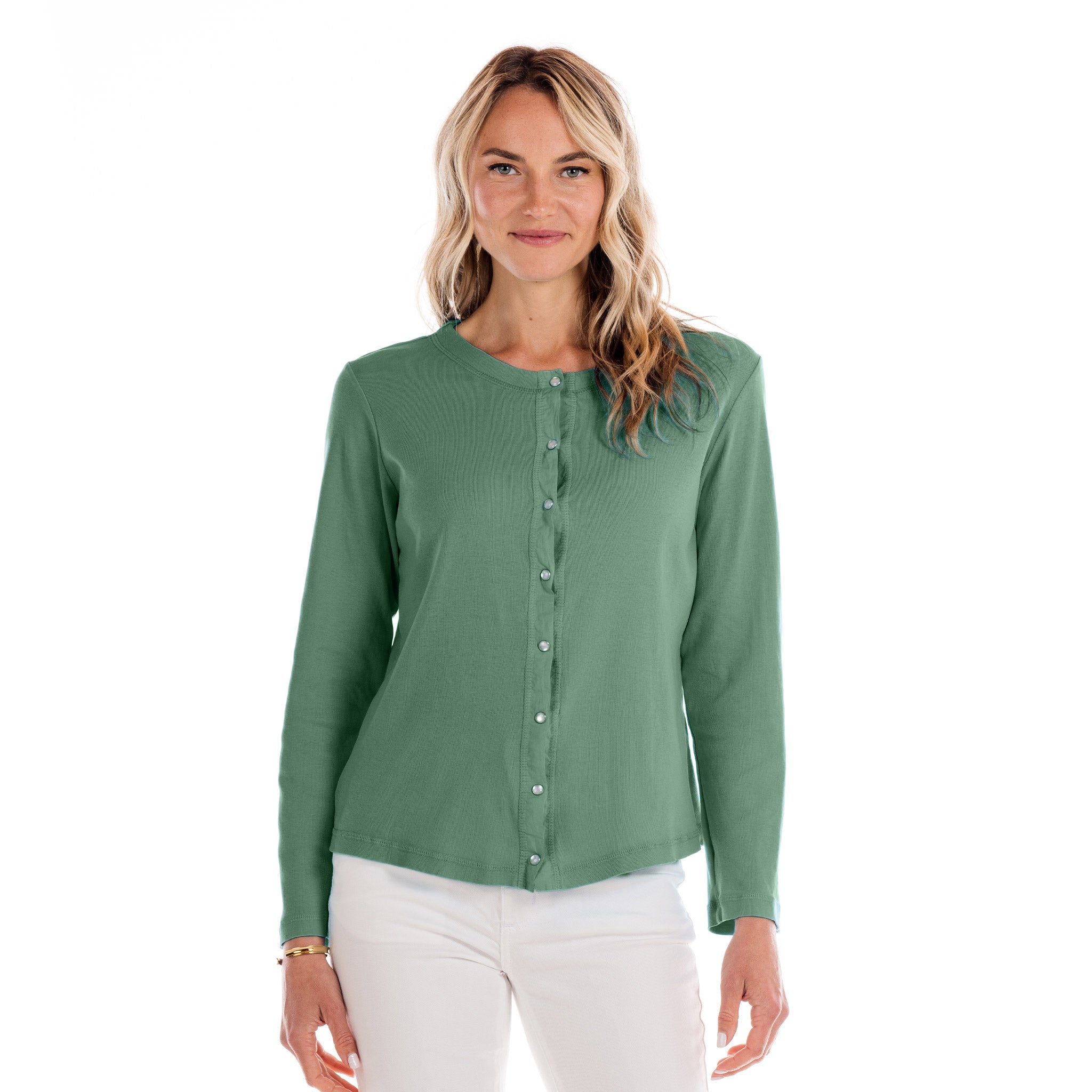 A woman with long blonde hair wears the Baby Rib Cardigan Lagoon by Fresh Produce Clothes, a green button-up sweater made from soft cotton rib, paired with white pants. She stands smiling against a plain white background.