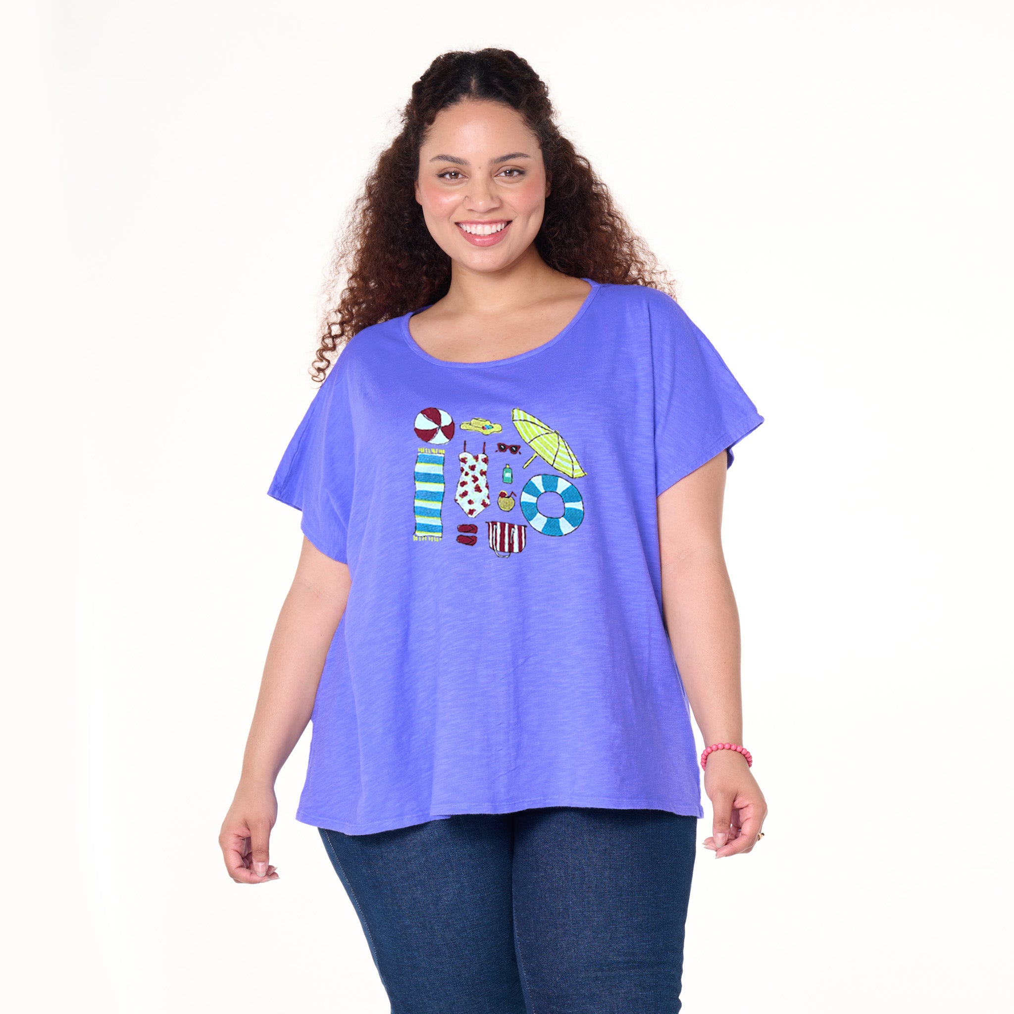 A smiling woman with curly hair wears the Fresh Produce Clothes Beach Day Essentials Keepsake T | Plus Size—a loose-fitting, short-sleeve purple tee adorned with colorful summer-inspired designs—perfectly styled with blue jeans and set against a white background.