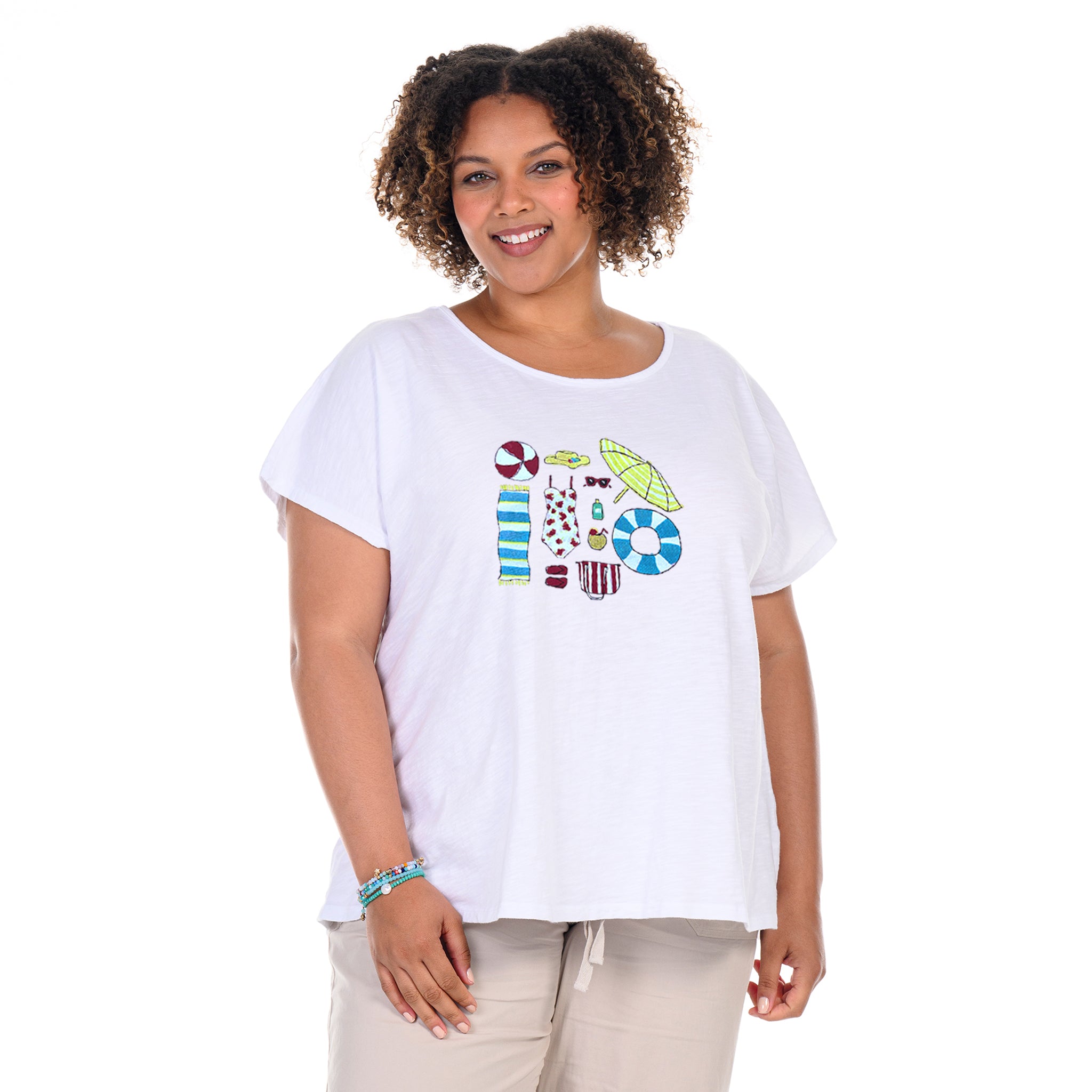 A woman with curly hair stands smiling against a plain white background, dressed casually in beige pants and the Fresh Produce Clothes Beach Day Essentials Keepsake T | Plus Size—featuring colorful beach-themed graphics for the perfect casual look.