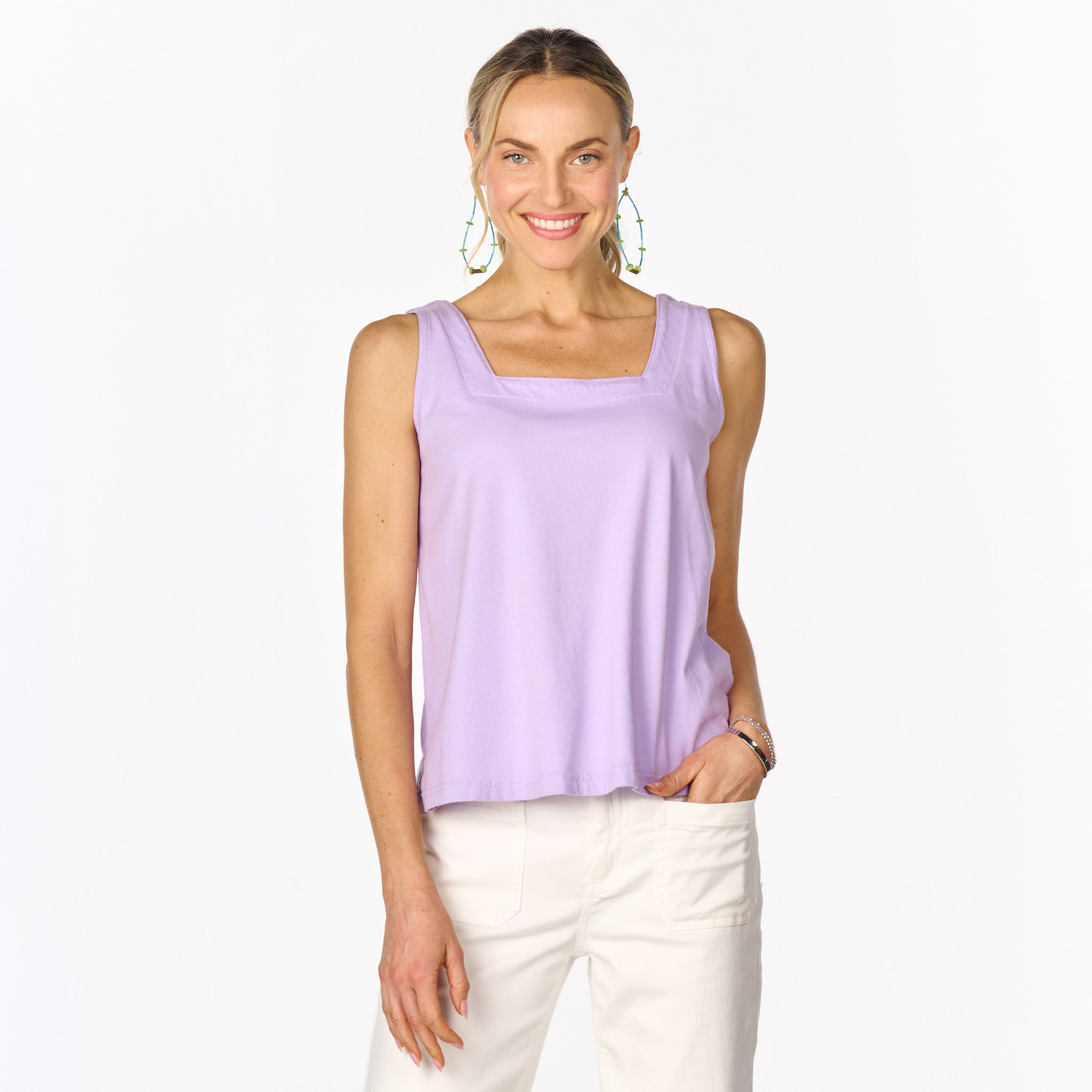 A woman smiles wearing the Fresh Produce Clothes Boxy Tank | Clearance, a bra-friendly, sleeveless lavender top with a square neckline and boxy silhouette, paired with white pants and large earrings as she stands against a plain white background.