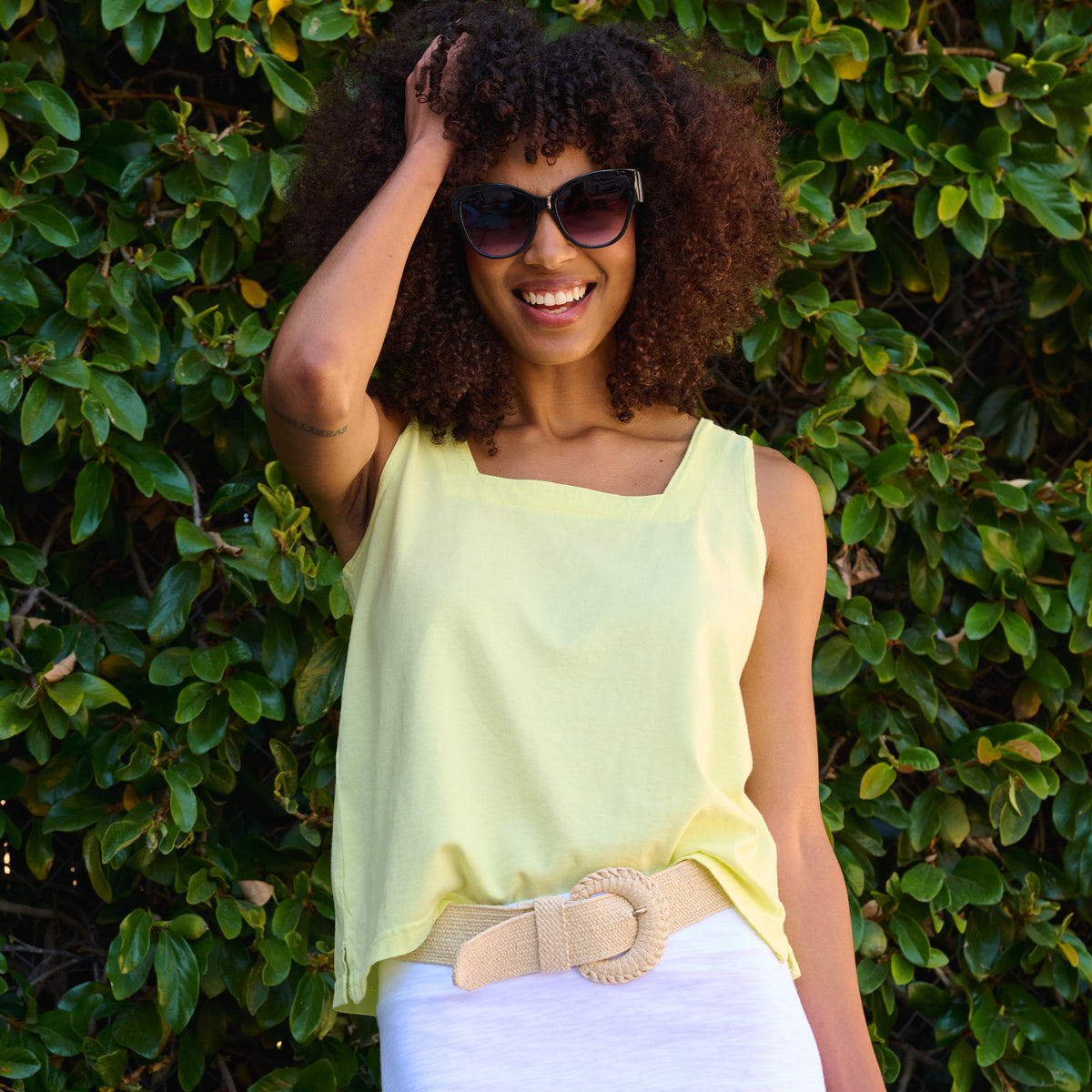 A woman with curly hair wearing sunglasses, the Boxy Tank in yellow by Fresh Produce Clothes, and a beige belt smiles while posing outdoors in front of green leafy bushes.