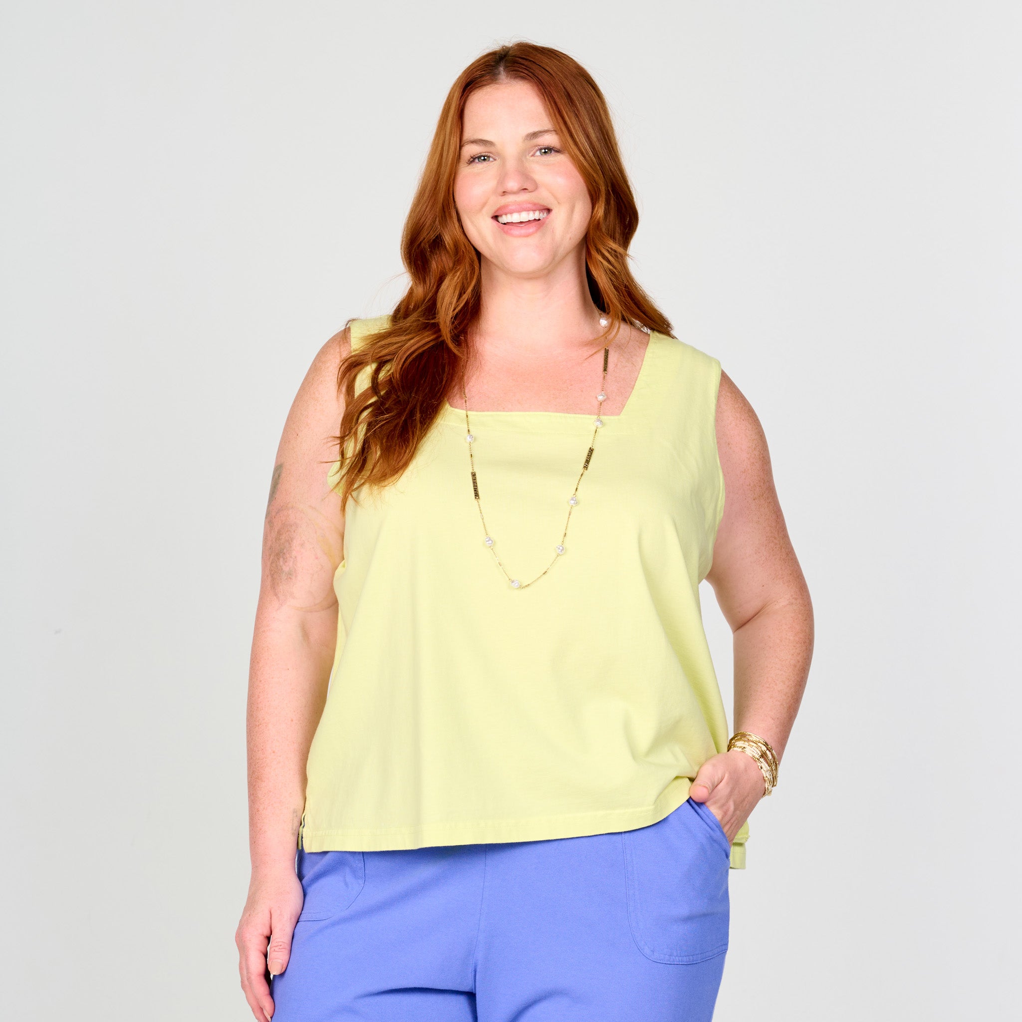 A smiling woman with long red hair models the Fresh Produce Clothes Boxy Tank | Plus Size | Clearance in light yellow. The bra-friendly, square neck tank is paired with blue pants, a long necklace, and a gold bracelet. She poses with her left hand in her pocket against a plain light background.