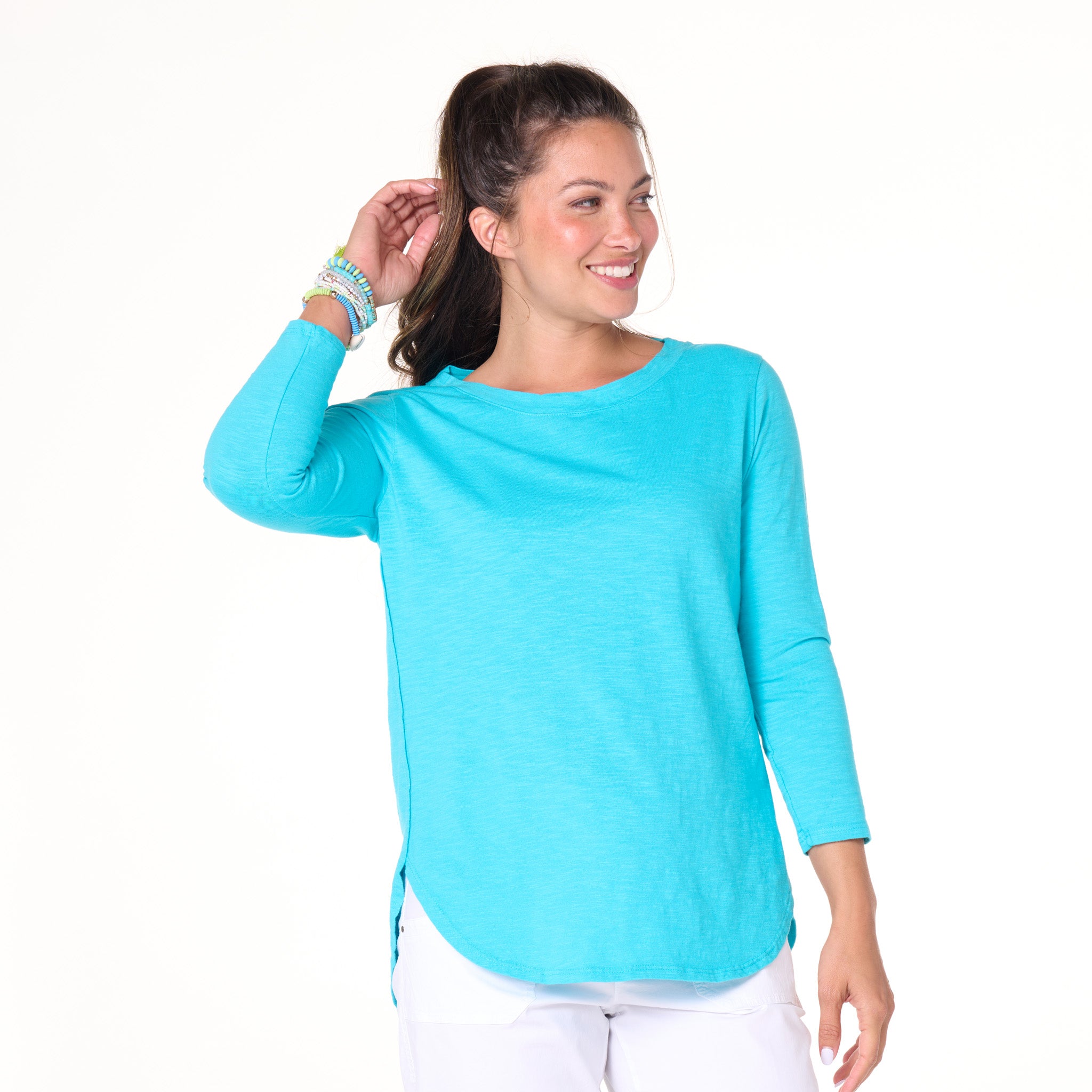 A woman stands smiling against a plain white background, wearing the Fresh Produce Clothes Catalina Top | Clearance in vibrant turquoise with a graceful scoop neckline, paired with classic white pants. One hand touches her ponytail while the other rests at her side.