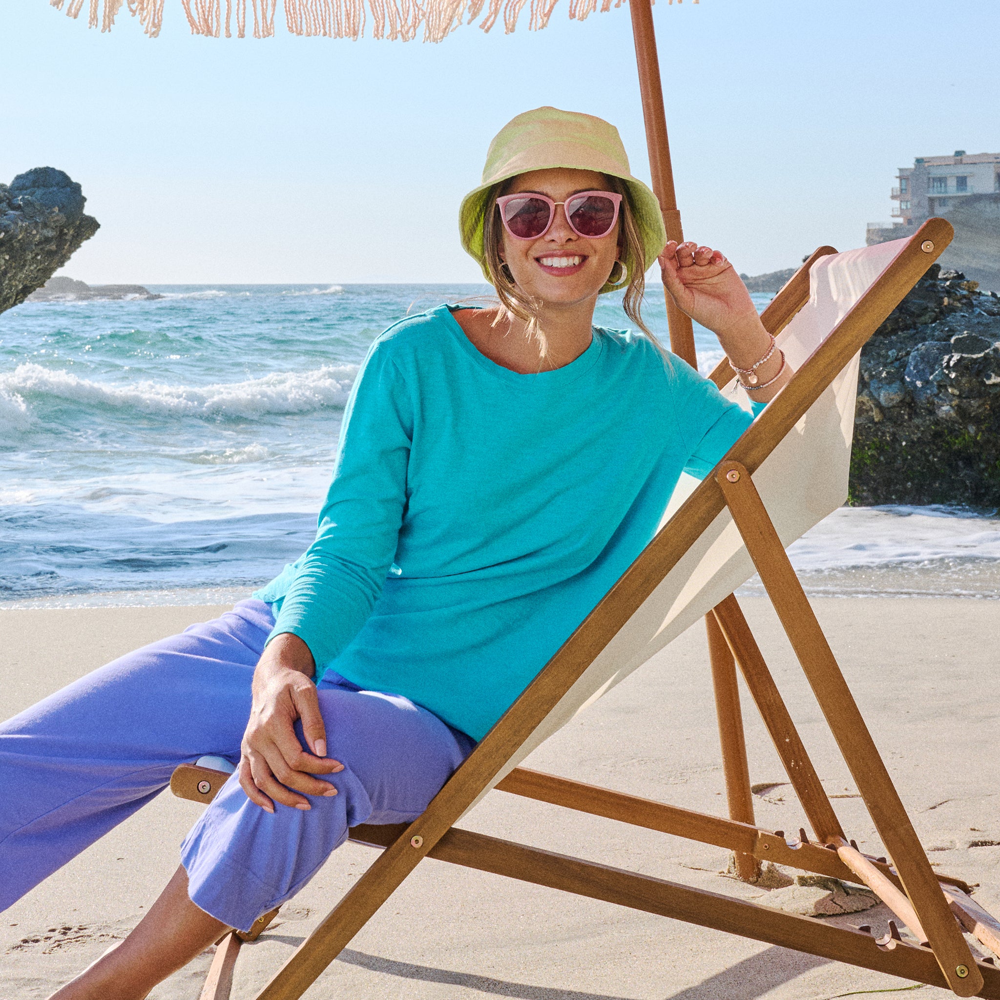 A woman in a green hat and sunglasses smiles while sitting on a wooden beach chair under an umbrella by the ocean, with waves and rocks in the background. She wears the Catalina Top from Fresh Produce Clothes, featuring a scoop neckline that adds breezy elegance to her seaside day.