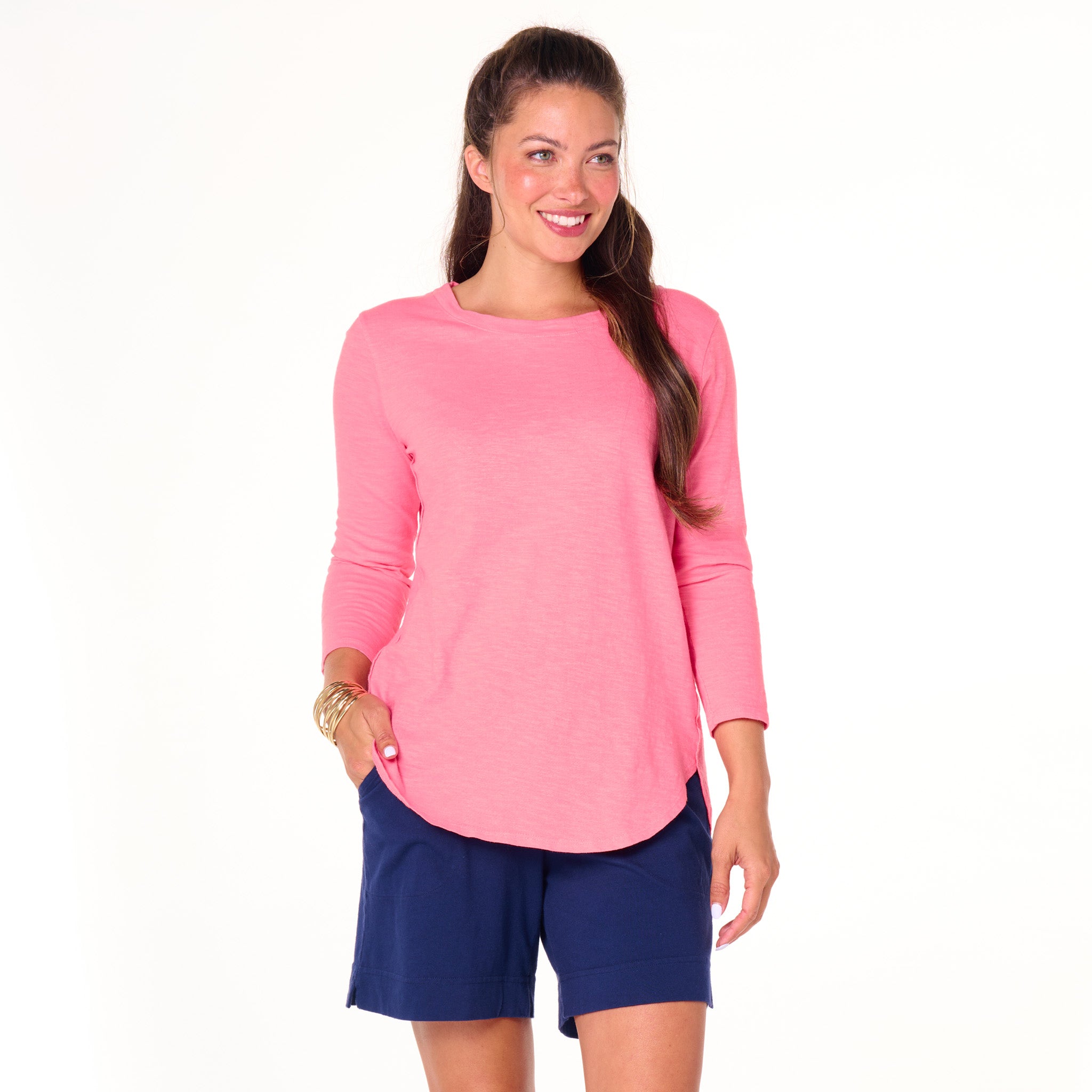A woman stands smiling with her hand in her pocket against a plain white background, wearing the Fresh Produce Clothes Catalina Top | Clearance in pink, featuring 3/4 sleeves and a flattering scoop neckline, perfectly paired with navy blue shorts.