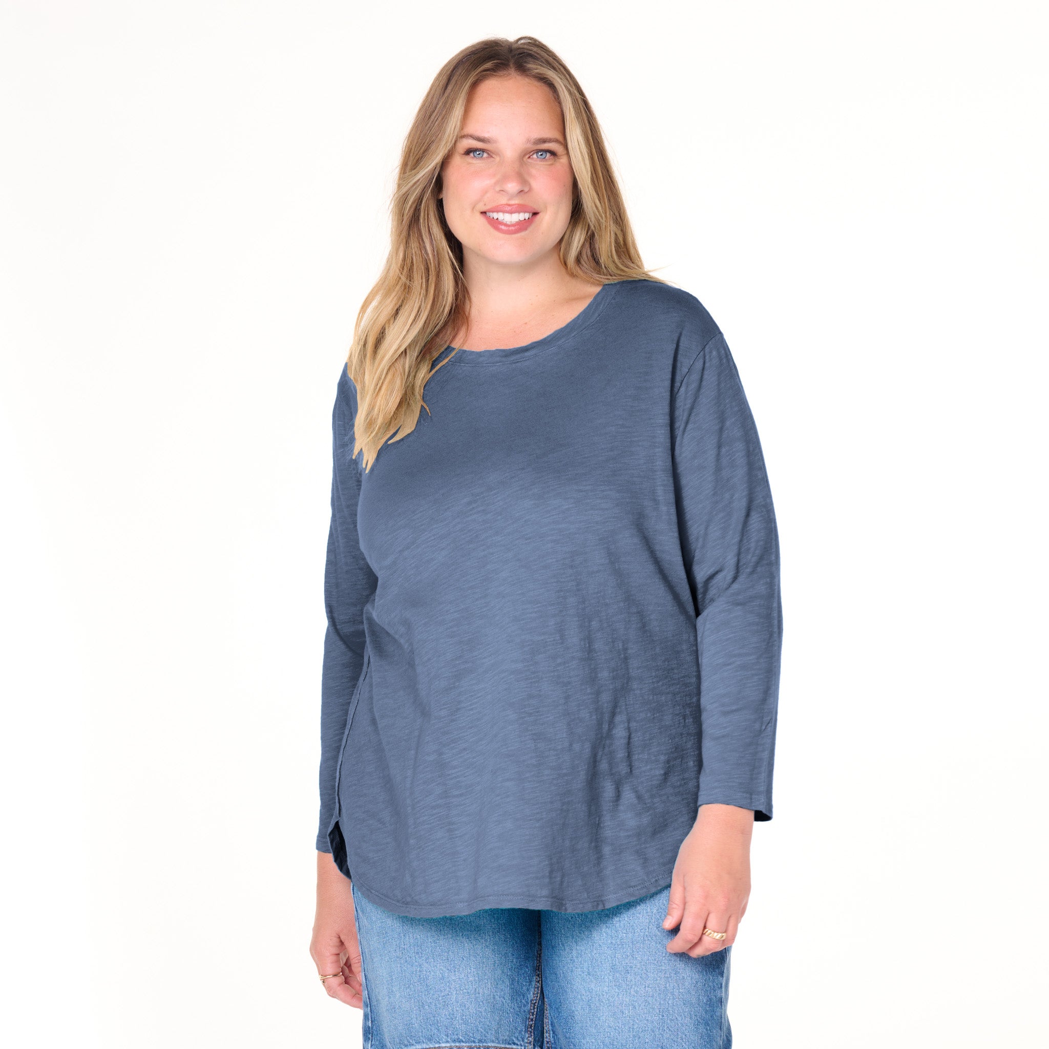 A woman with long blonde hair wears the Fresh Produce Clothes Catalina Top | Plus Size in slate grey, featuring 3/4 sleeves and a curved hemline, paired with light blue jeans. She smiles while standing against a plain white background.