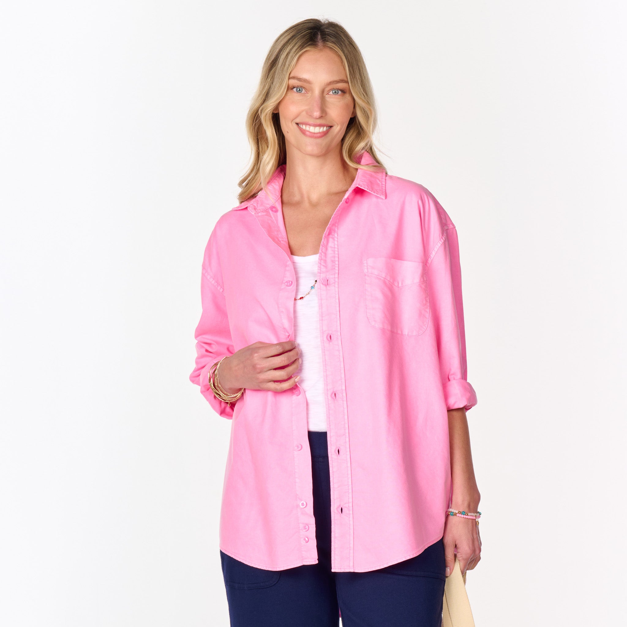 A woman with long blonde hair smiles while wearing the Fresh Produce Clothes Classic Oxford Shirt in bright pink (Clearance), styled over a white top and dark blue pants, standing against a plain white background.
