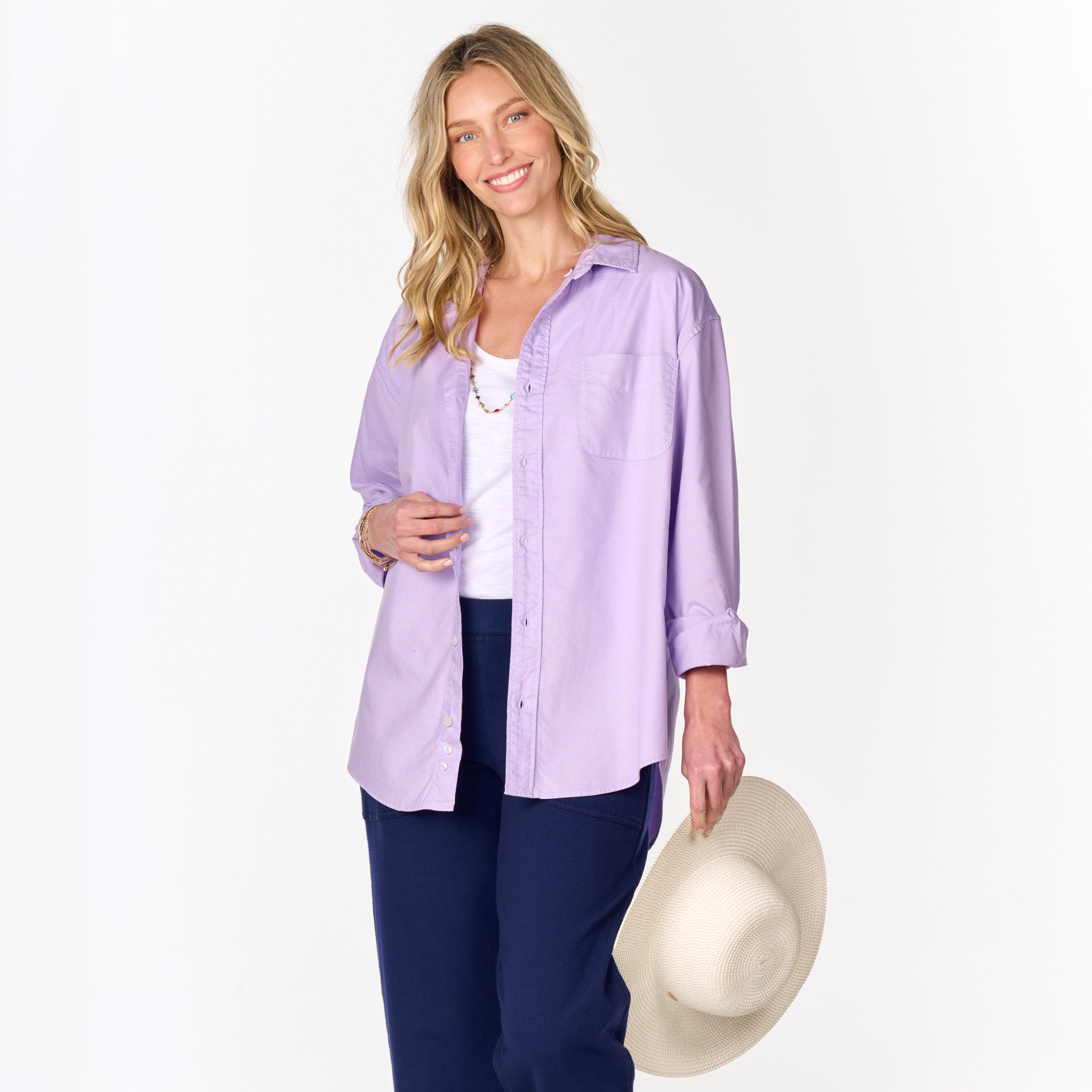 A woman with long blonde hair wears the Fresh Produce Clothes Classic Oxford Shirt—a relaxed fit, light purple button-up styled as an oversized layer over a white top and dark pants. She smiles and holds a wide-brimmed straw hat in her left hand, with a plain white background behind her. This item is featured in Clearance.