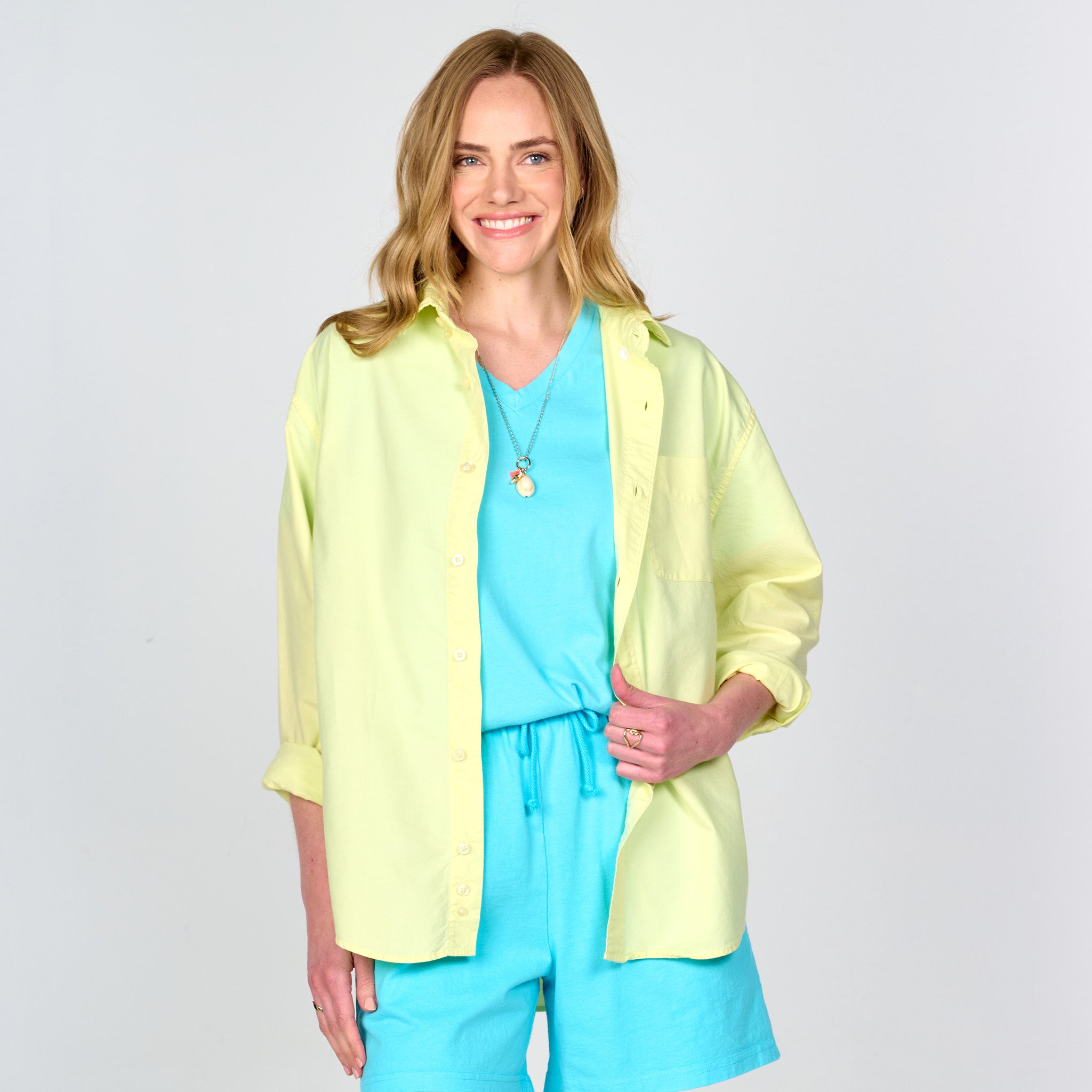 A woman with long blonde hair smiles, wearing the Fresh Produce Clothes Classic Oxford Shirt | Clearance in light yellow over a turquoise top and shorts, accessorized with a necklace, standing against a plain light background.