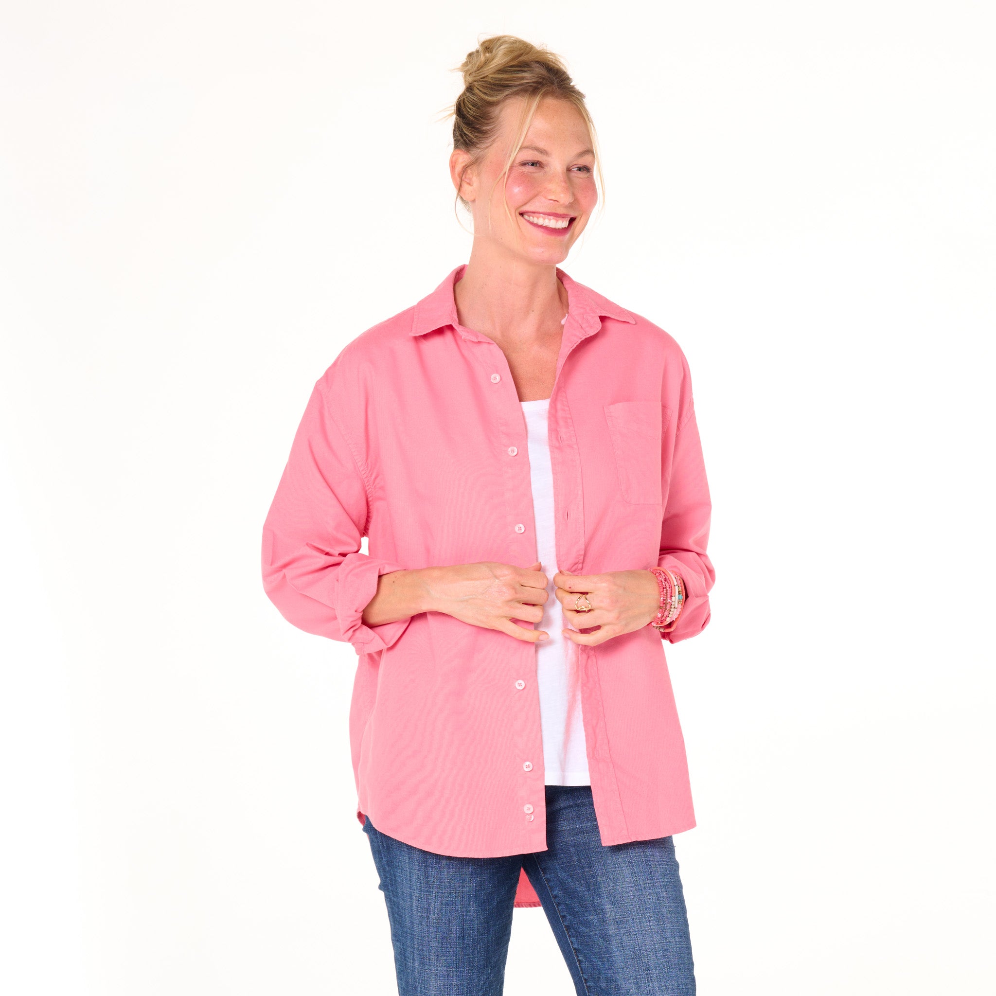 A smiling woman models the Fresh Produce Clothes Classic Oxford Shirt in a relaxed fit pink style, layered over a white top and blue jeans, standing against a plain white background.