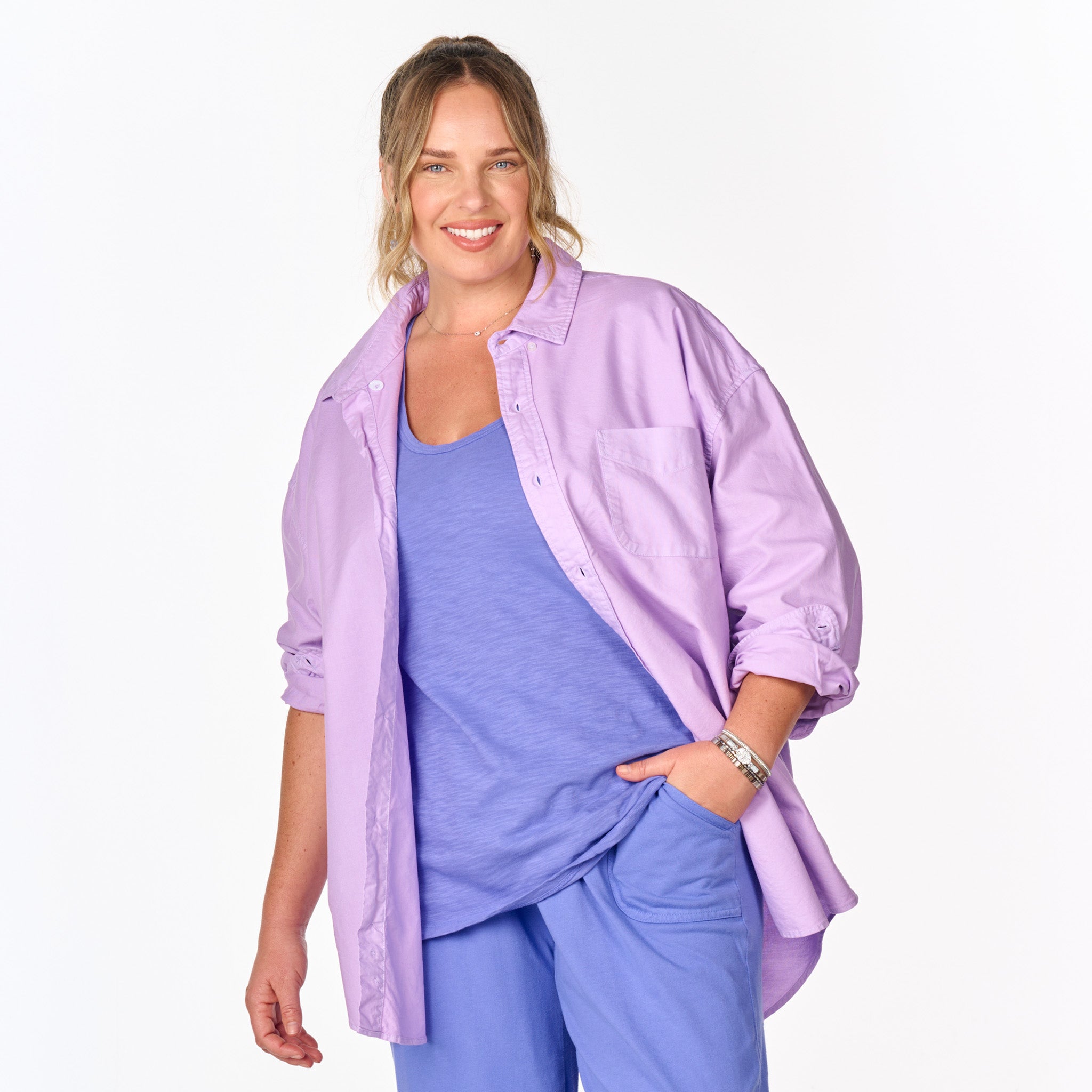 A smiling woman with light brown hair in a loose ponytail is wearing the Fresh Produce Clothes Classic Oxford Shirt (Plus Size, Clearance) in lavender over a blue top and blue pants, standing against a plain white background.