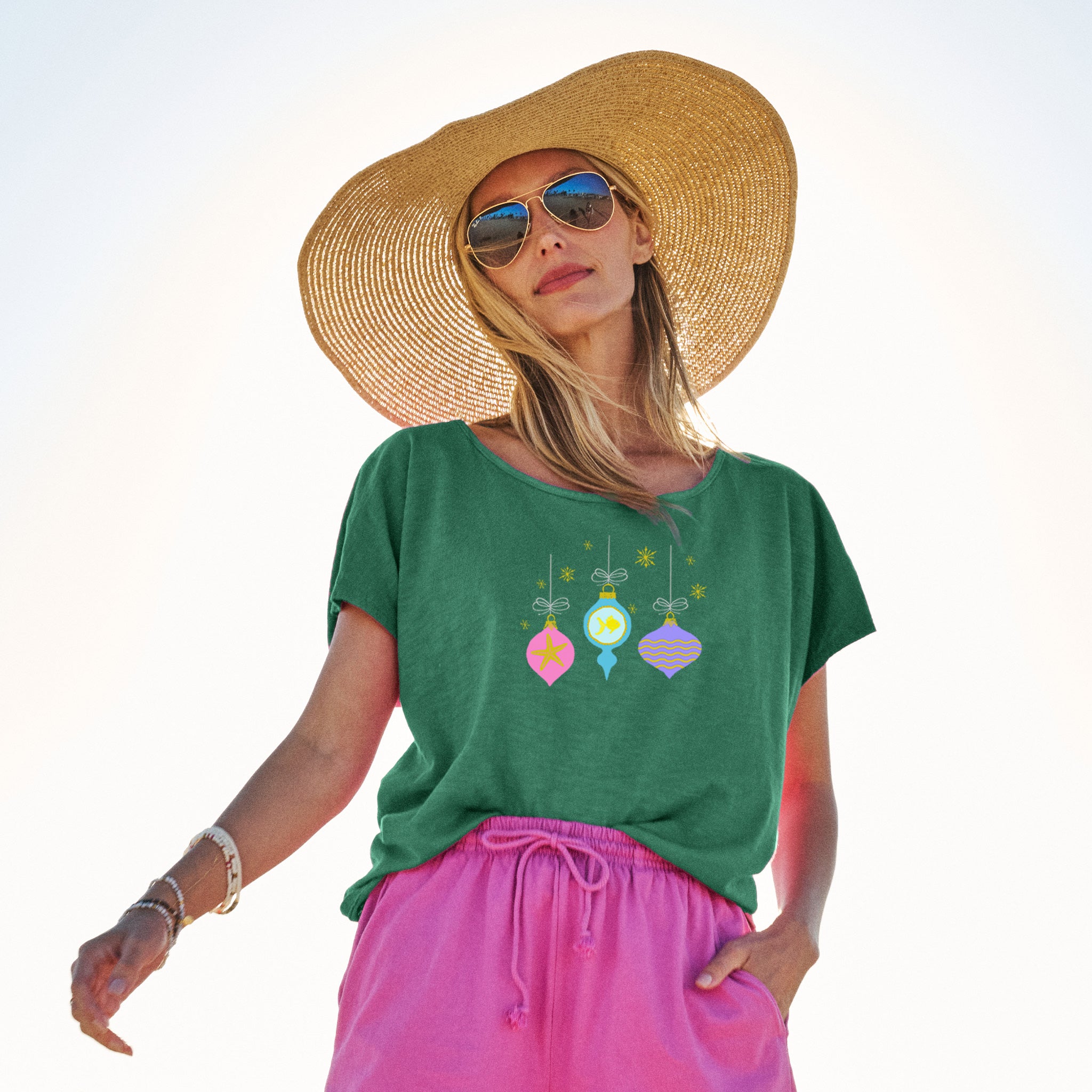 A woman wearing a large straw hat, sunglasses, and the Fresh Produce Clothes Coastal Ornaments Keepsake T in green, paired with pink shorts, stands outdoors against a bright sky. She looks confidently at the camera—her style radiates effortless holiday spirit.