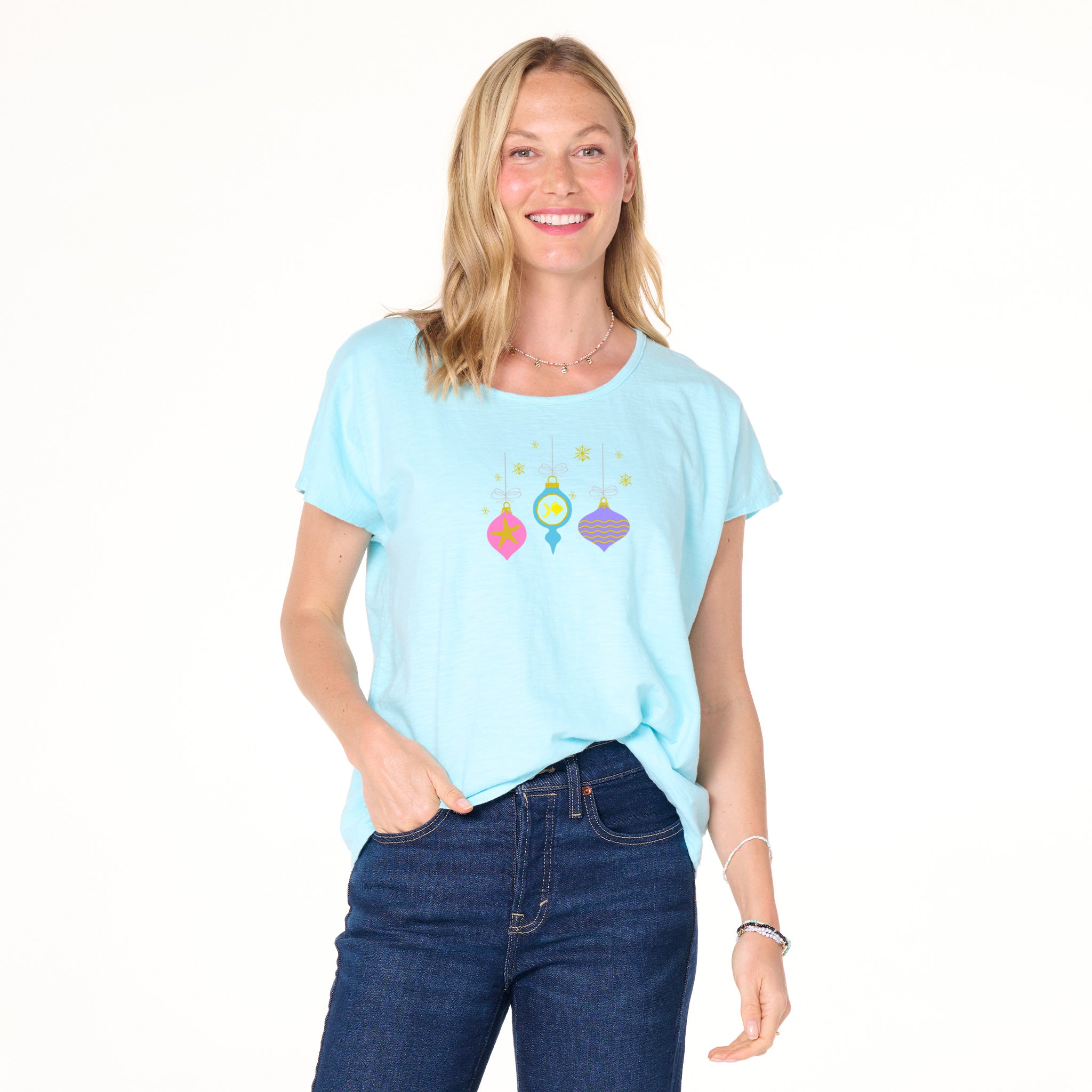 A smiling woman with blonde hair wears a light blue Fresh Produce Clothes Coastal Ornaments Keepsake T featuring festive ornament graphics, paired with dark blue jeans. Standing against a white background with one hand in her pocket, she perfectly captures the relaxed holiday spirit.