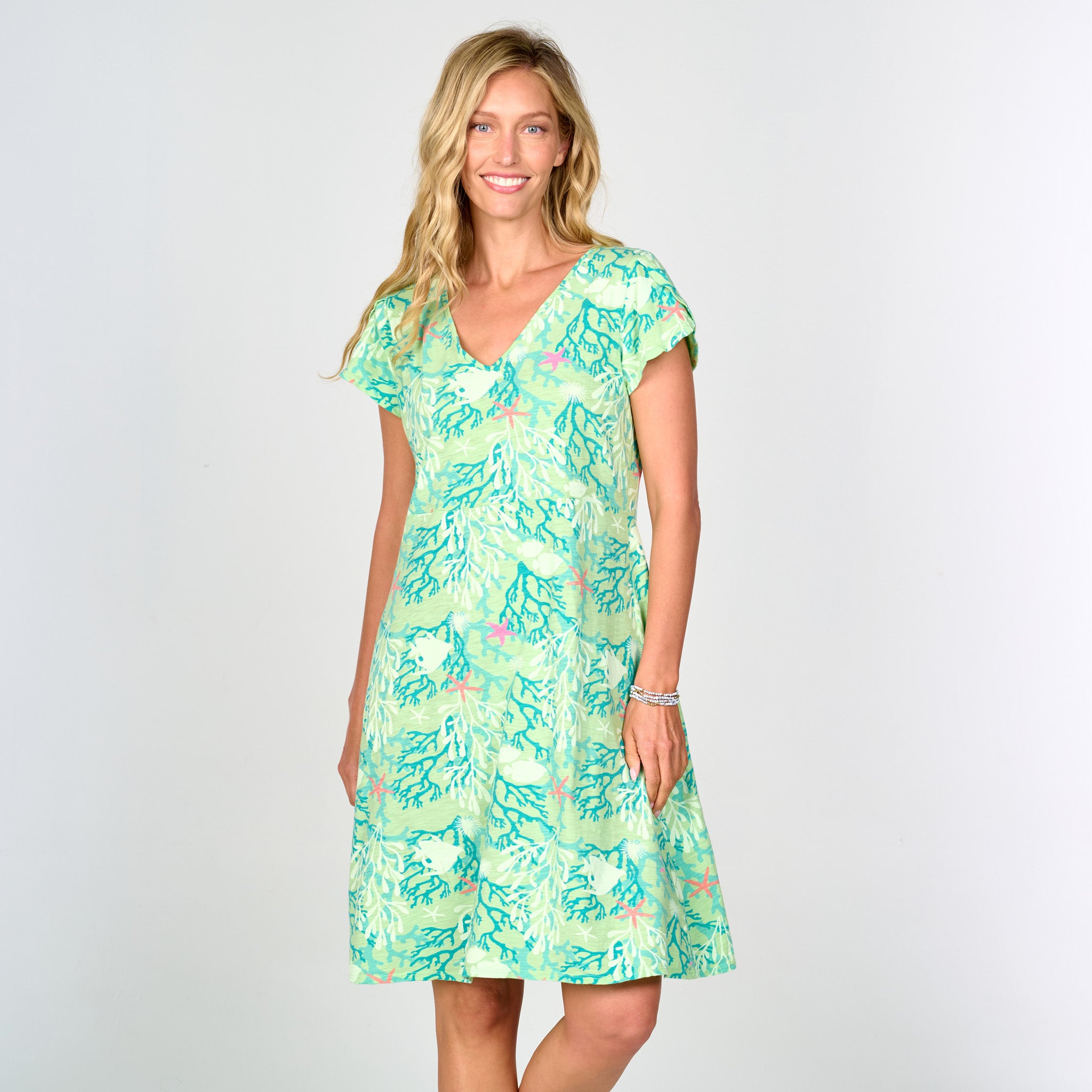 A woman with long blonde hair smiles while standing against a plain white background, wearing the Fresh Produce Clothes Coral Reef Amelia Dress in light green, which features a leafy pattern, tulip sleeves, and an elegant empire waist.